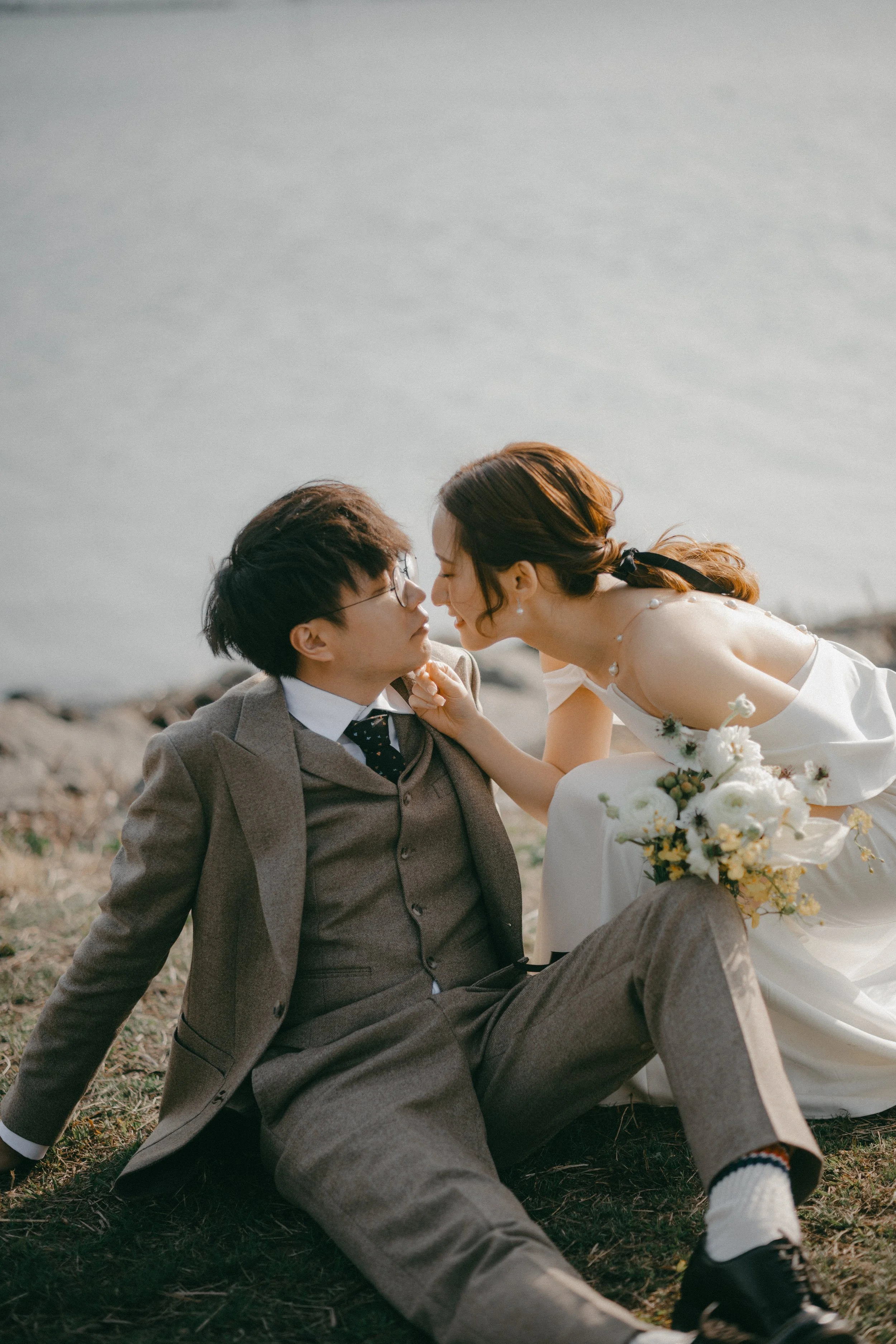 A couple dressed in wedding attire sitting on grass near water, with the woman leaning towards the man.