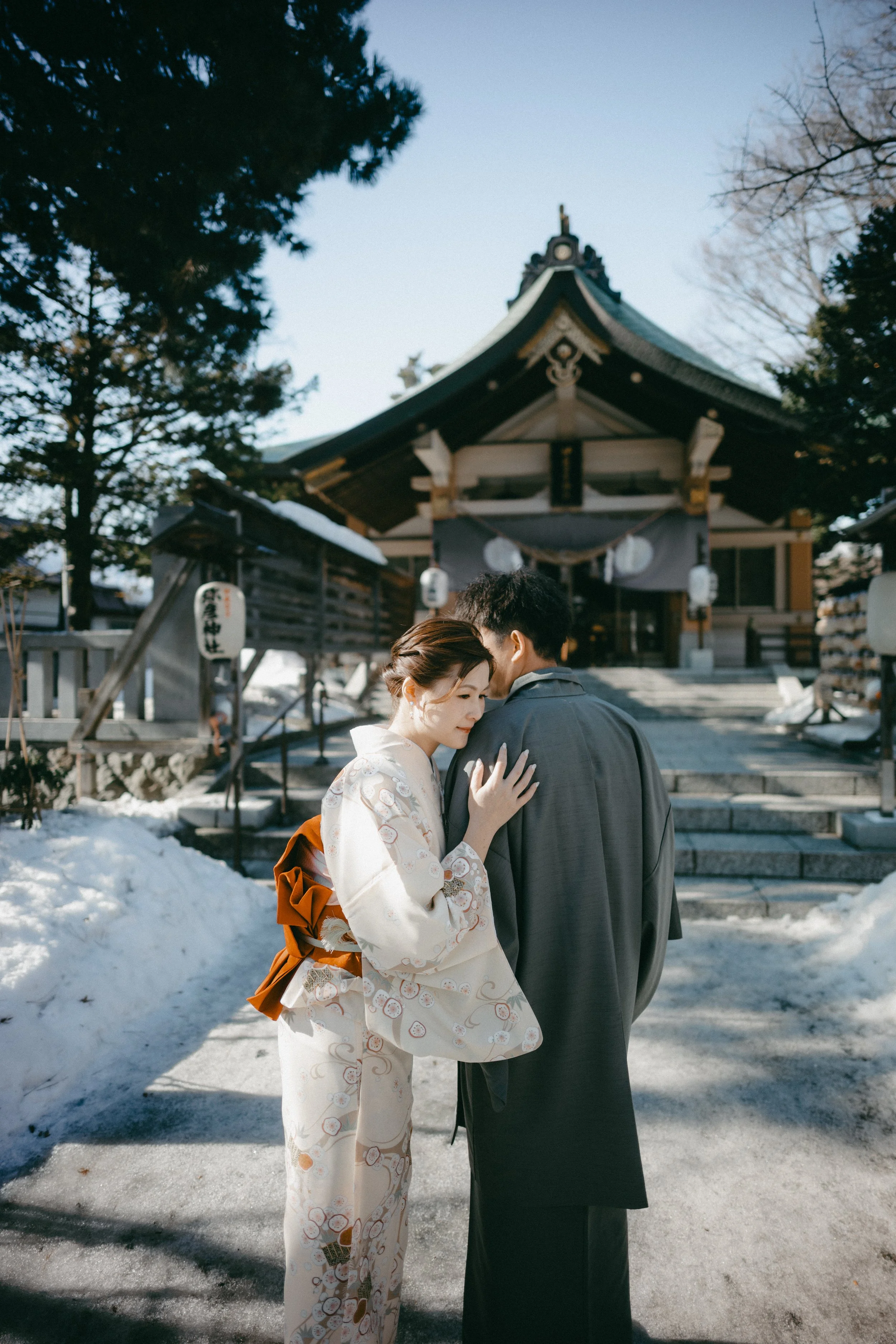 A couple dressed in traditional Japanese wedding attire sharing an intimate moment in front of a Shinto shrine in winter.