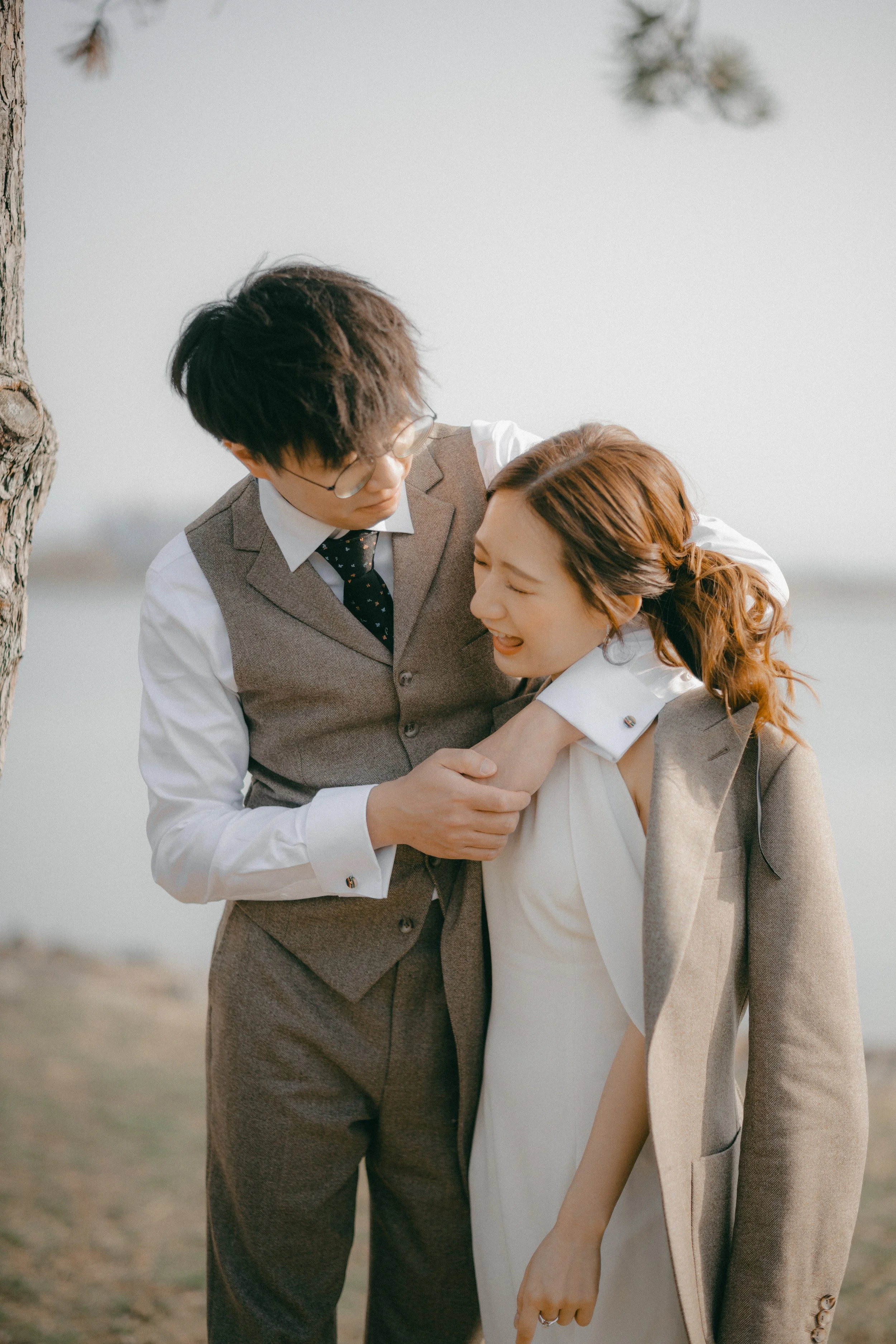 A man and a woman laughing and playing together outdoors near a tree by a body of water. The man is wearing glasses, a vest over a shirt, and a tie, while the woman has red hair styled in loose waves and is wearing a white dress with a beige blazer.