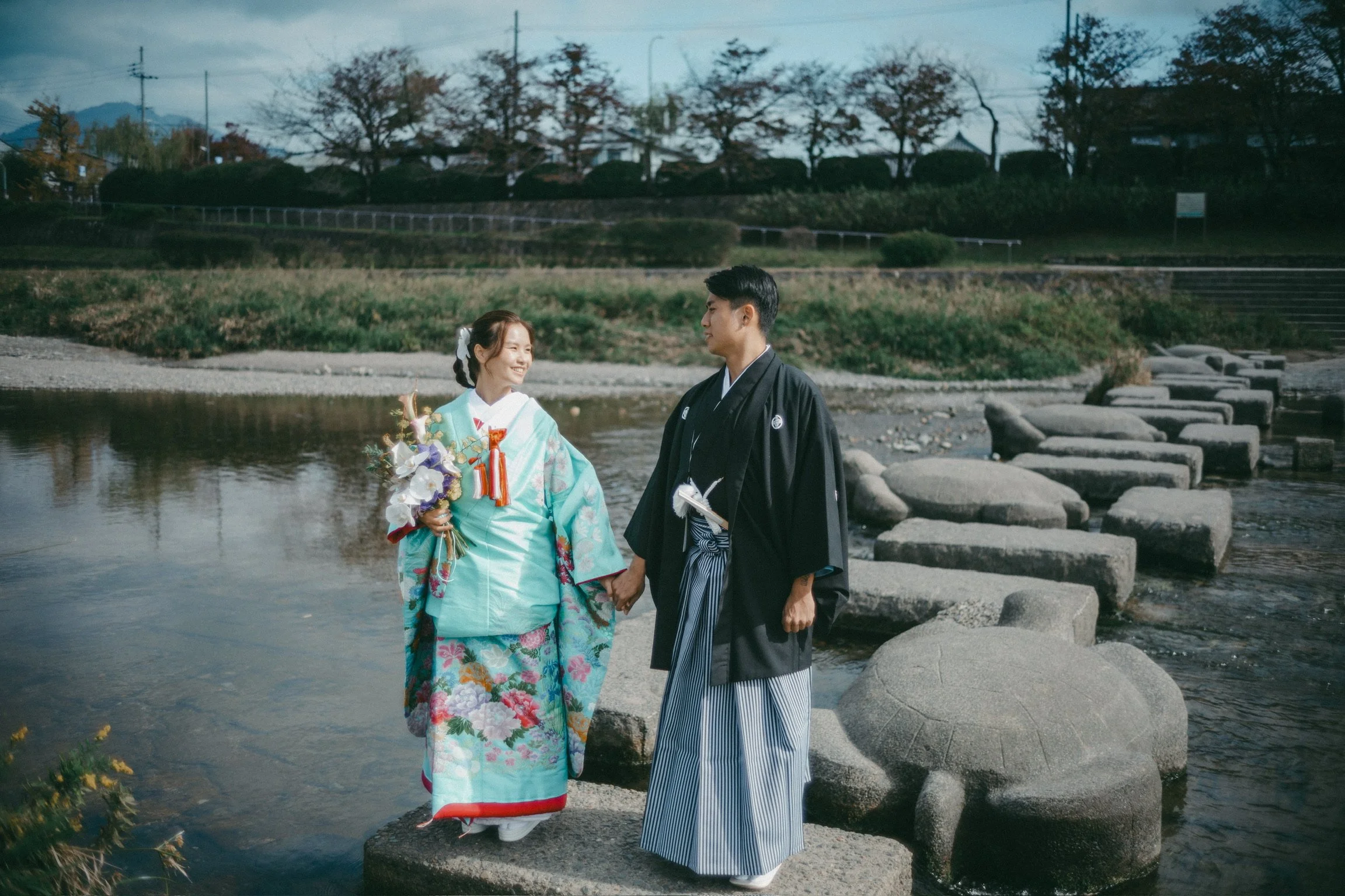 A Japanese couple in traditional attire holding hands on stepping stones in a river, smiling at each other.