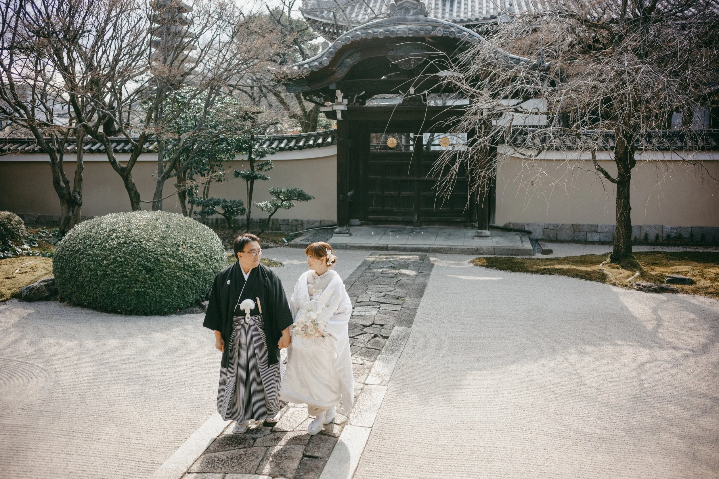A bride and groom holding hands walking on a stone path in a traditional Japanese garden with trees, bushes, and a wooden gate in the background.