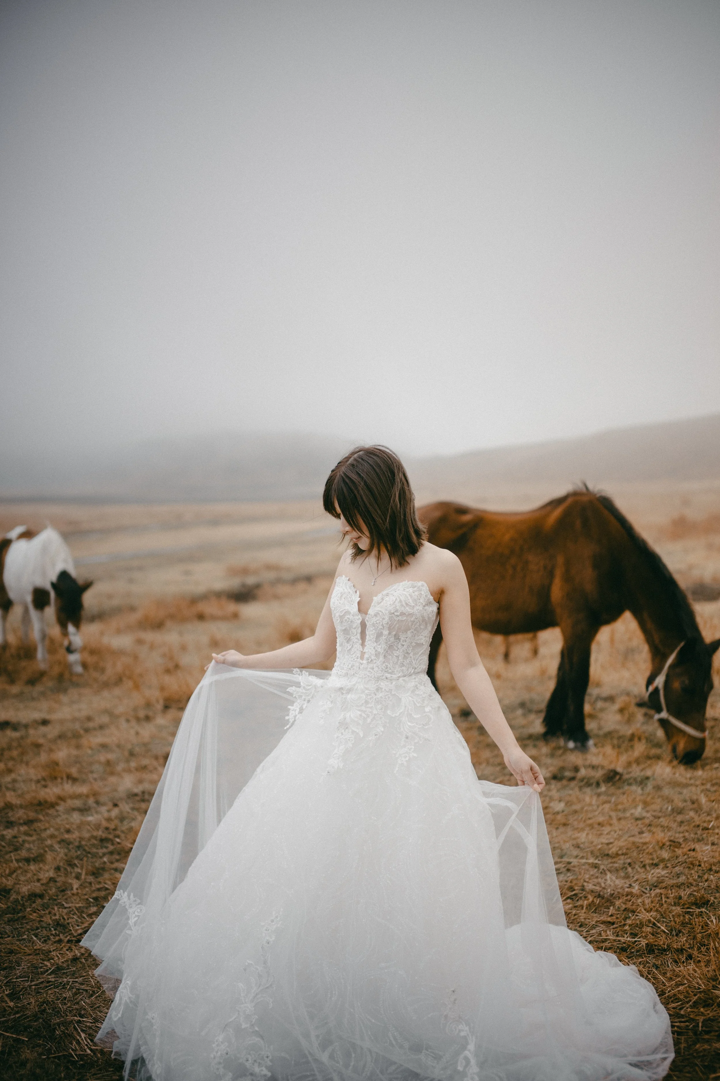 A woman in a white wedding dress standing in a field with two horses grazing nearby under a cloudy sky.