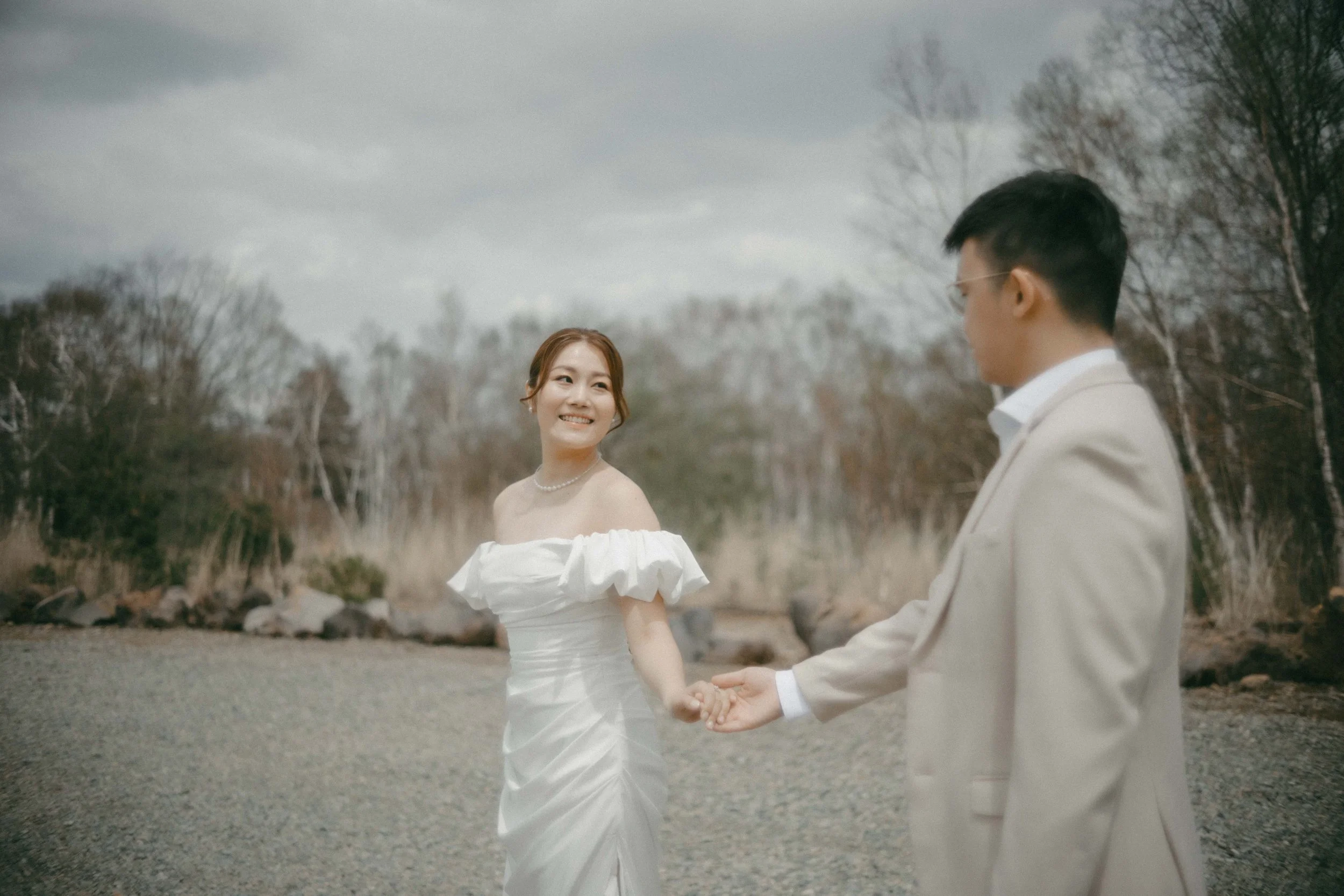 A bride and groom holding hands outdoors on a cloudy day, with trees in the background.