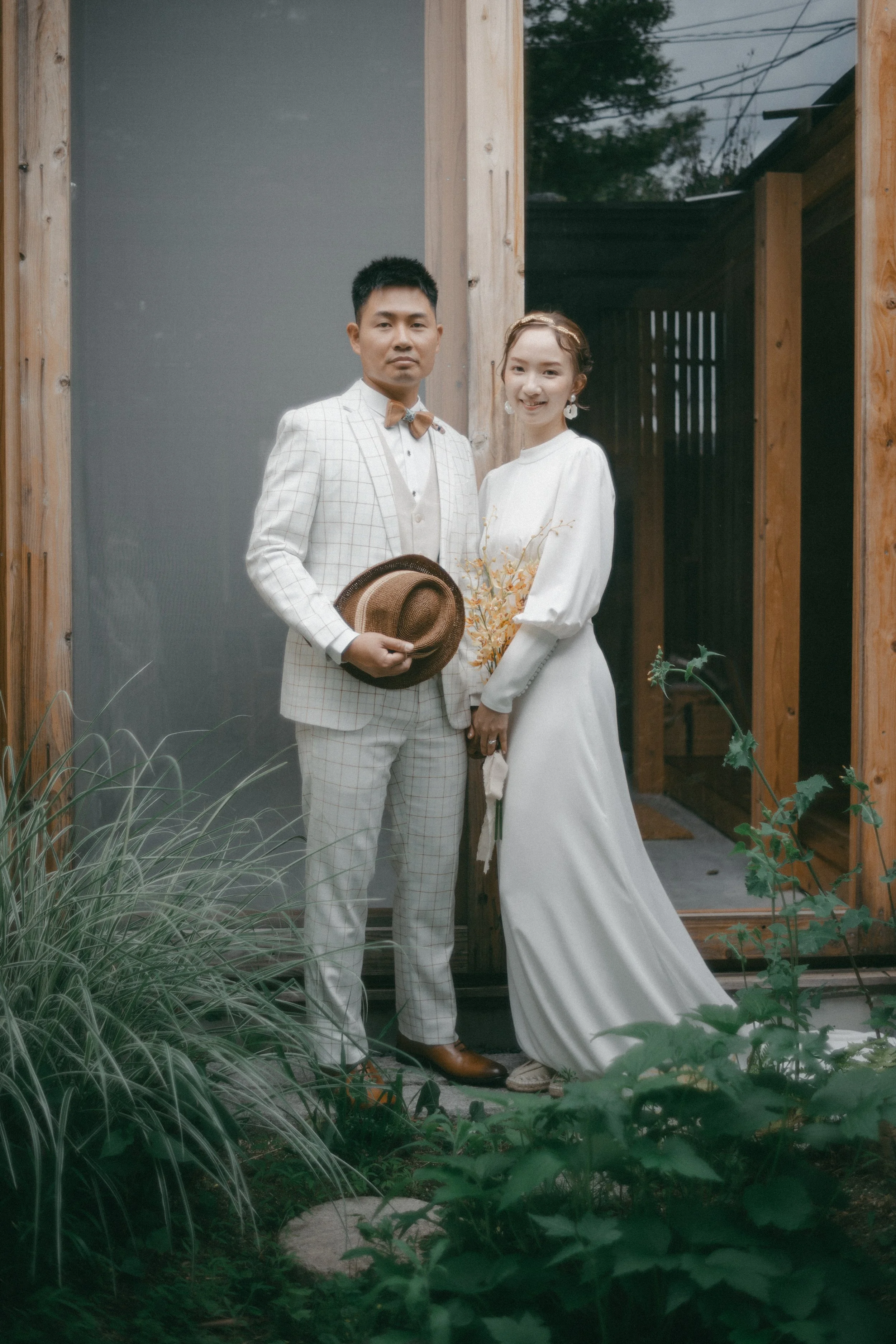 A man and woman dressed in wedding attire standing outside near a wooden door with greenery in the foreground.