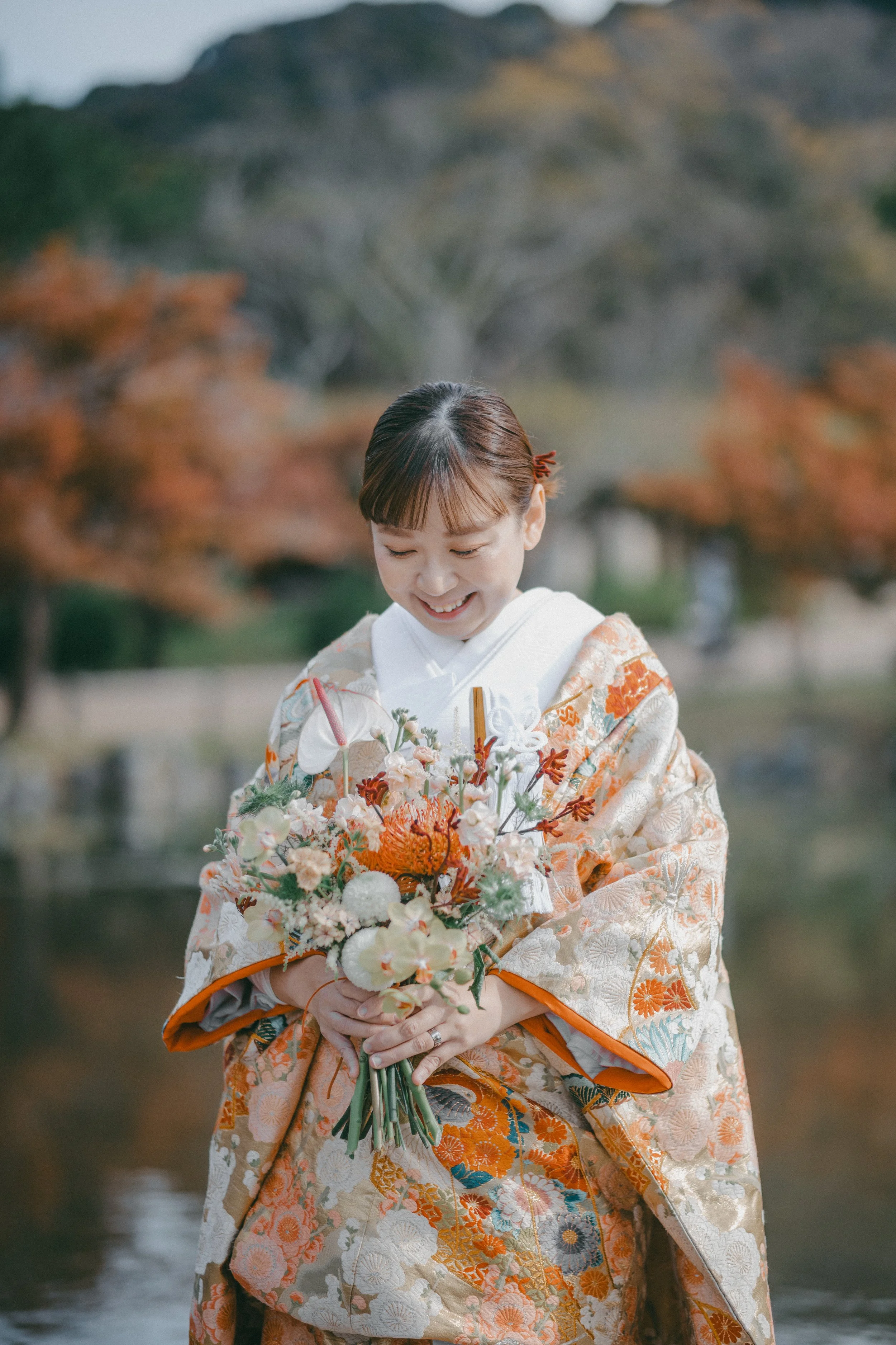 A woman in a traditional Japanese kimono holding a bouquet of flowers outdoors, with autumn foliage in the background.