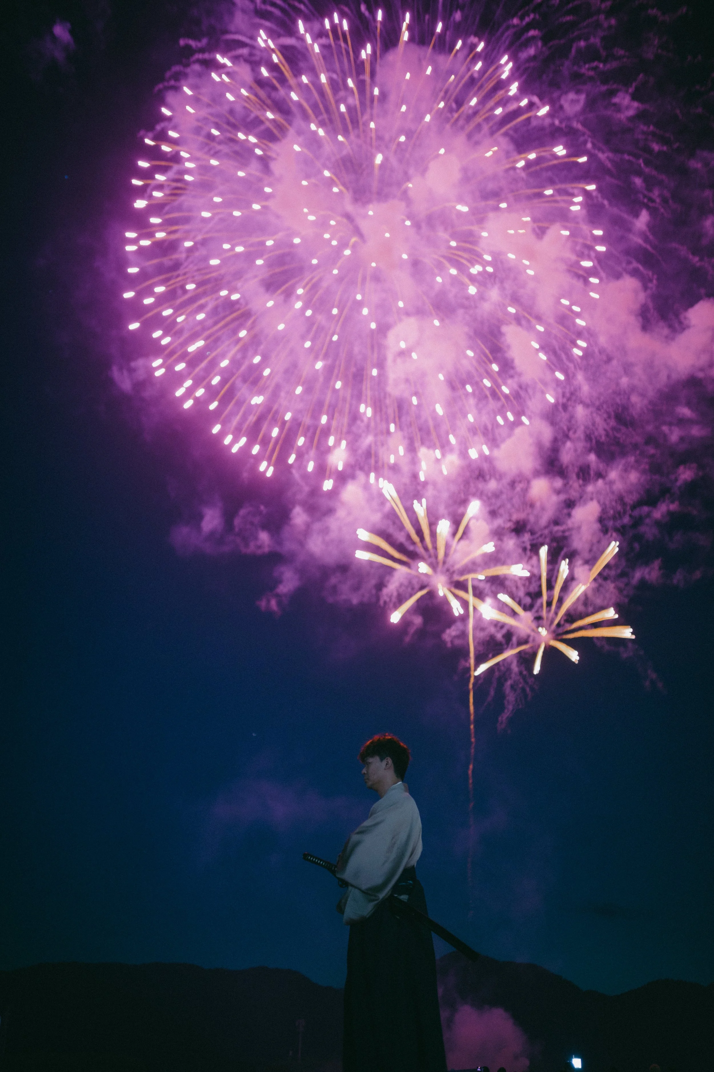 A person dressed in traditional Japanese attire holding a katana, standing under a dark night sky with colorful fireworks exploding overhead.