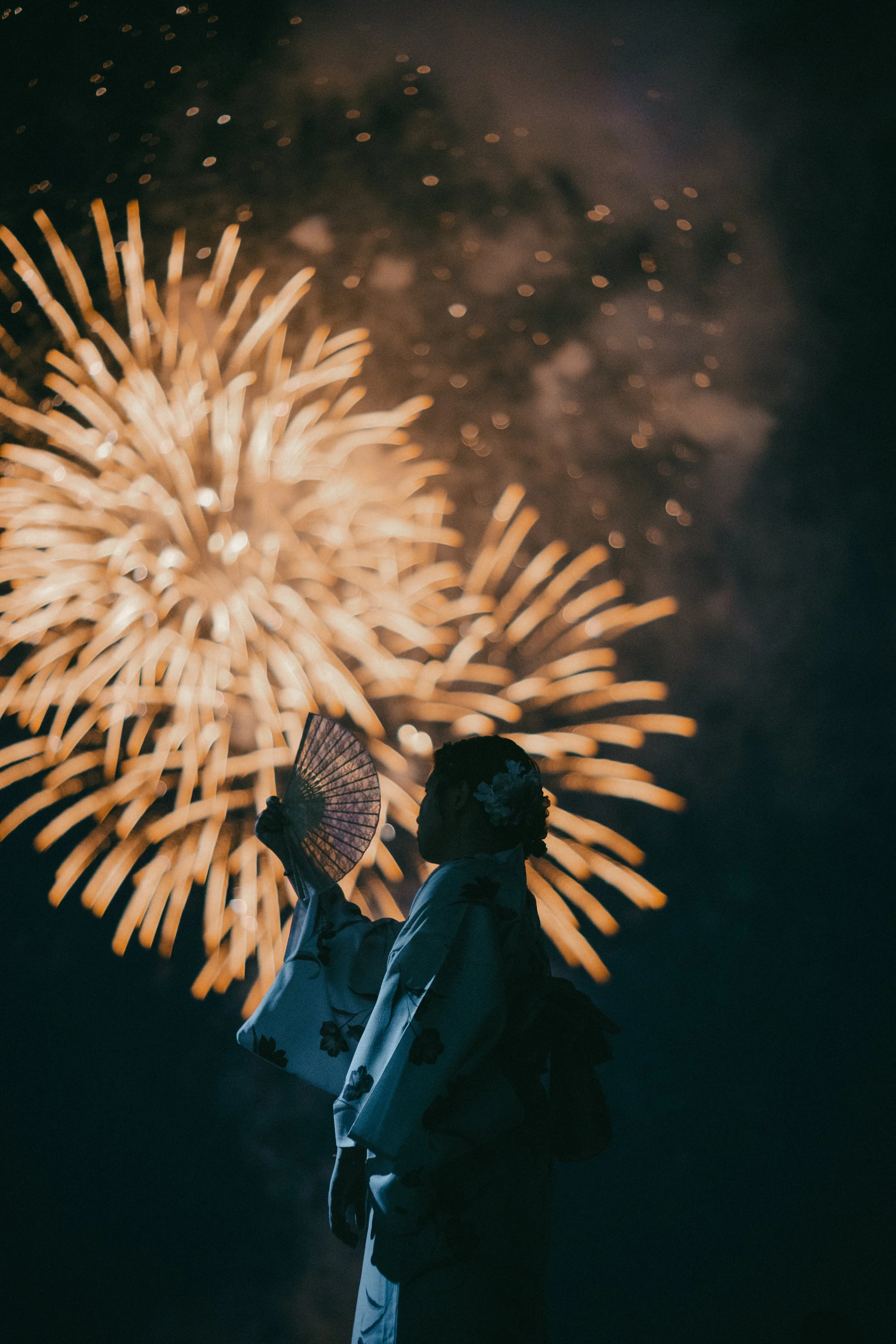 A silhouette of a woman holding a paper fan, standing in front of colorful fireworks at night.