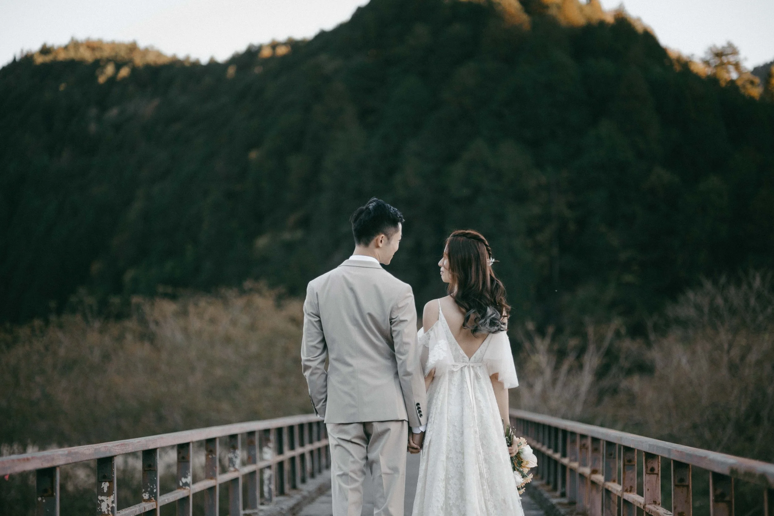A bride and groom holding hands on a bridge outdoors, with mountains and trees in the background, during sunset.
