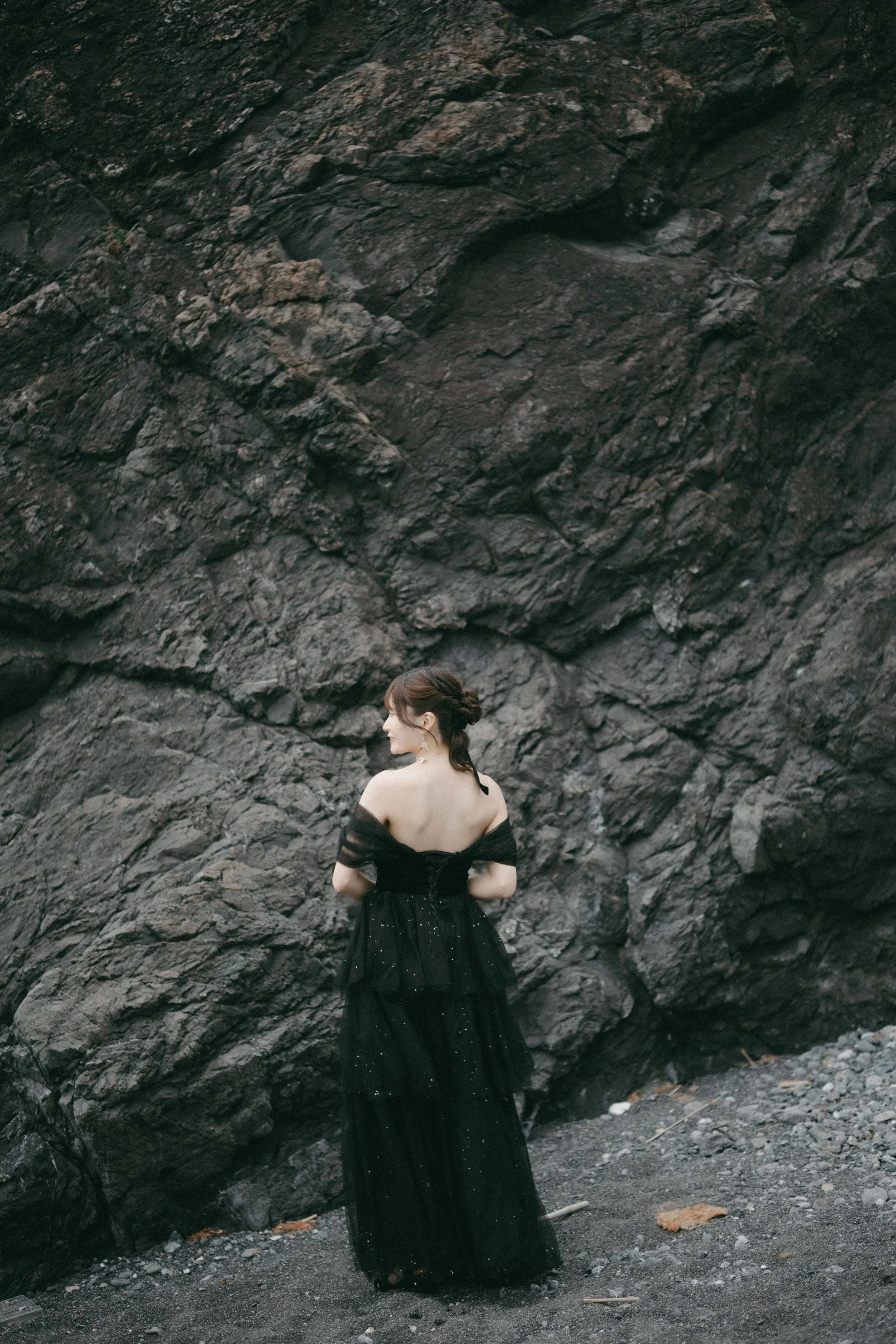 A woman in a black, off-shoulder, floor-length dress standing in front of a large rocky cliff, with her back turned and head slightly to the side, at an outdoor location.