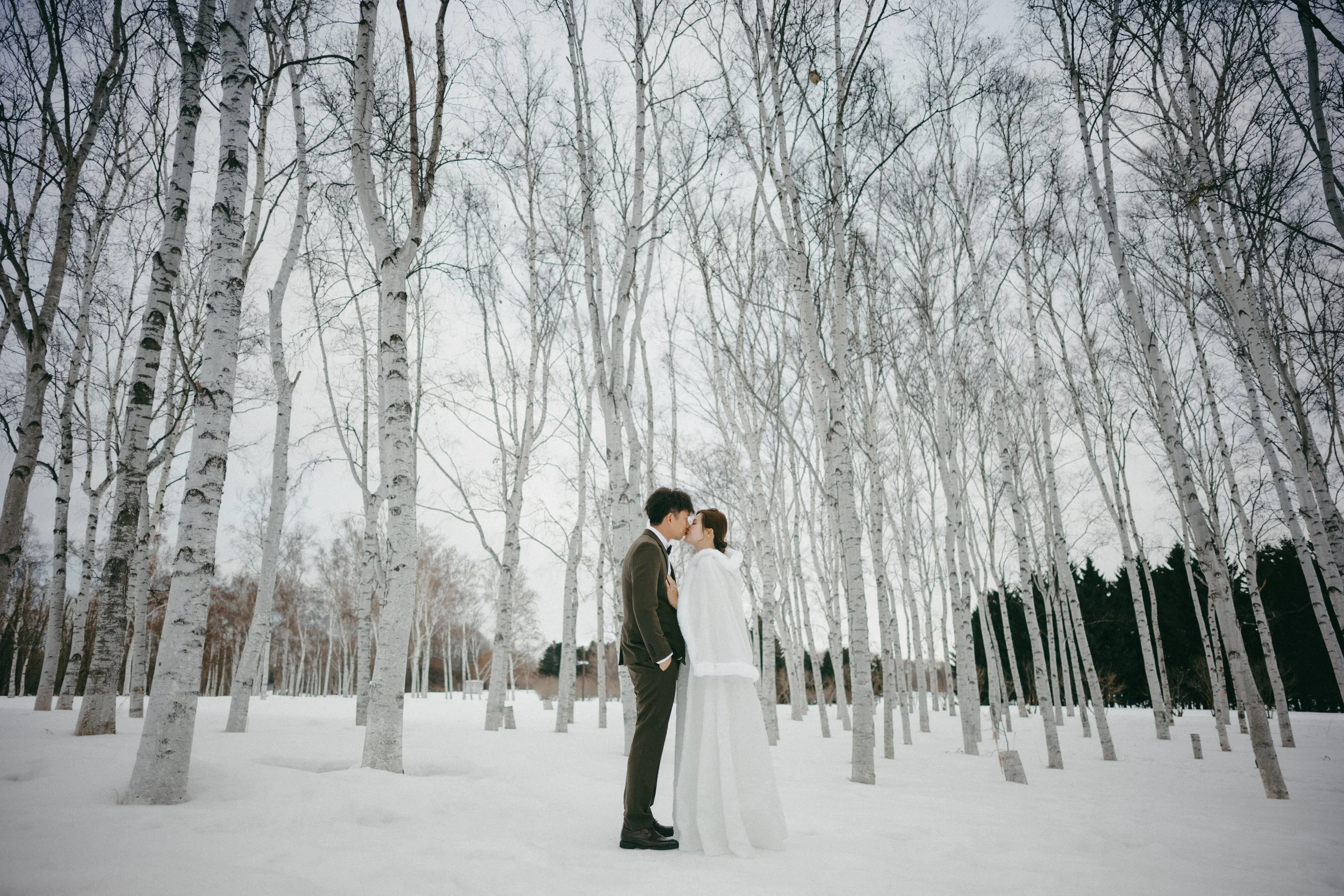 A couple dressed in wedding attire kissing in a snowy forest with tall, leafless trees.