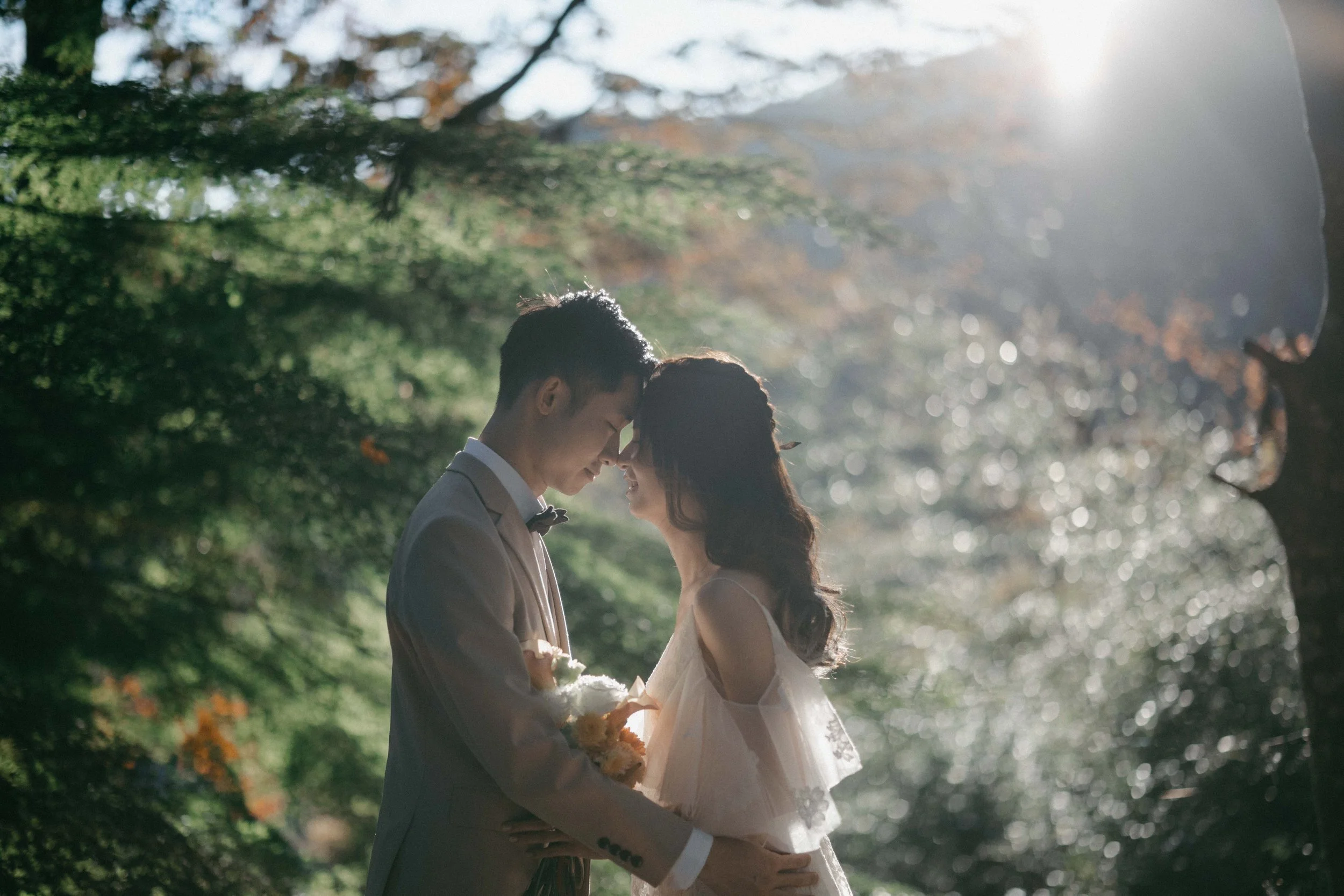 A bride and groom sharing an intimate moment with foreheads touching in a sunlit outdoor setting surrounded by trees.