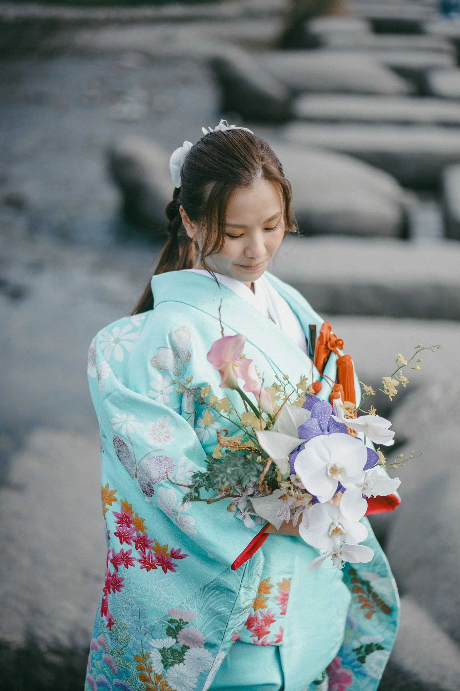 A young woman wearing a traditional turquoise Japanese kimono decorated with floral and butterfly patterns, holding a bouquet of white orchids and pink flowers, standing near a river with rocks.