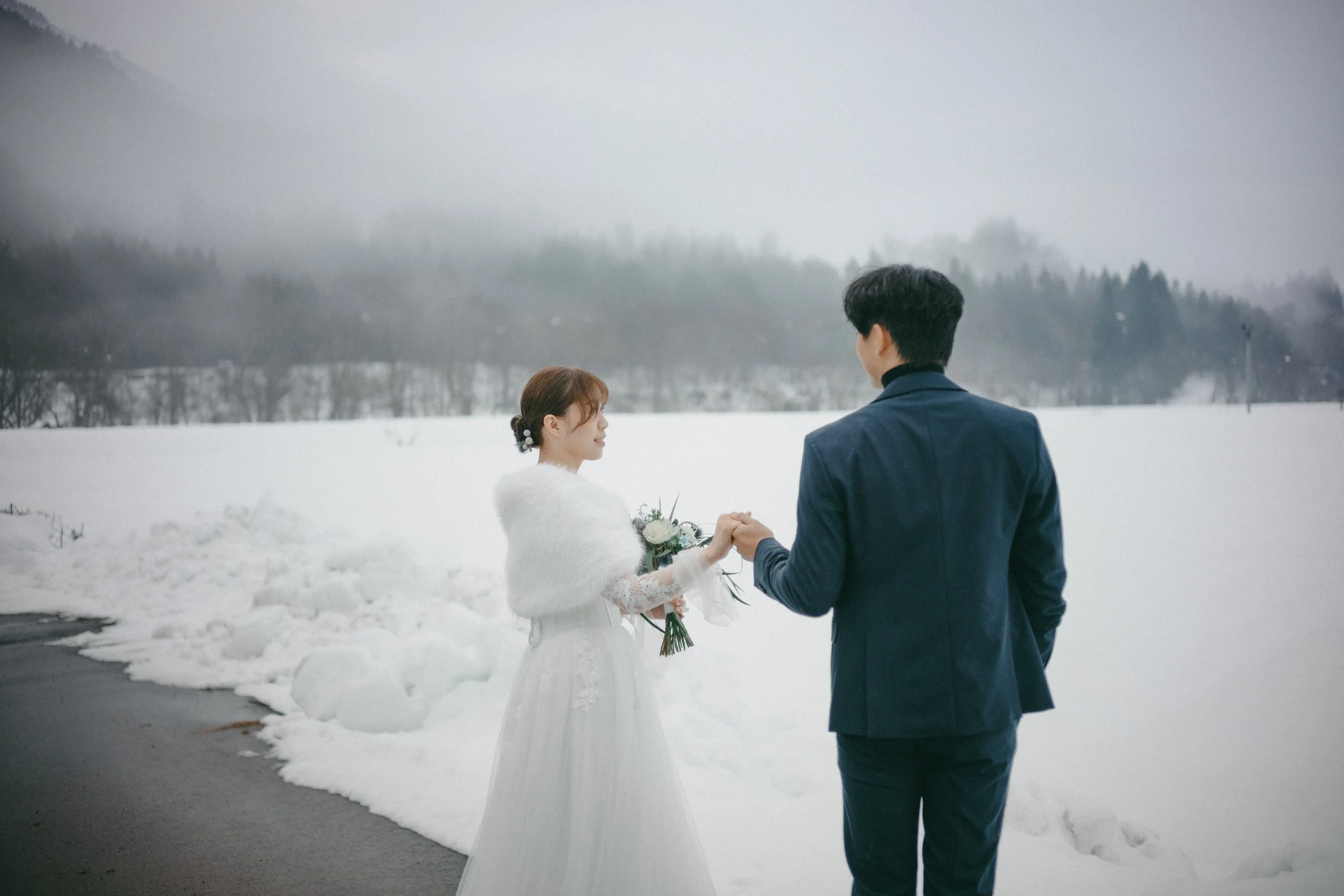 A bride and groom on a snowy landscape, holding hands, with the groom facing away from the camera and the bride smiling, holding a bouquet, surrounded by snow-covered ground and mountain in the background.