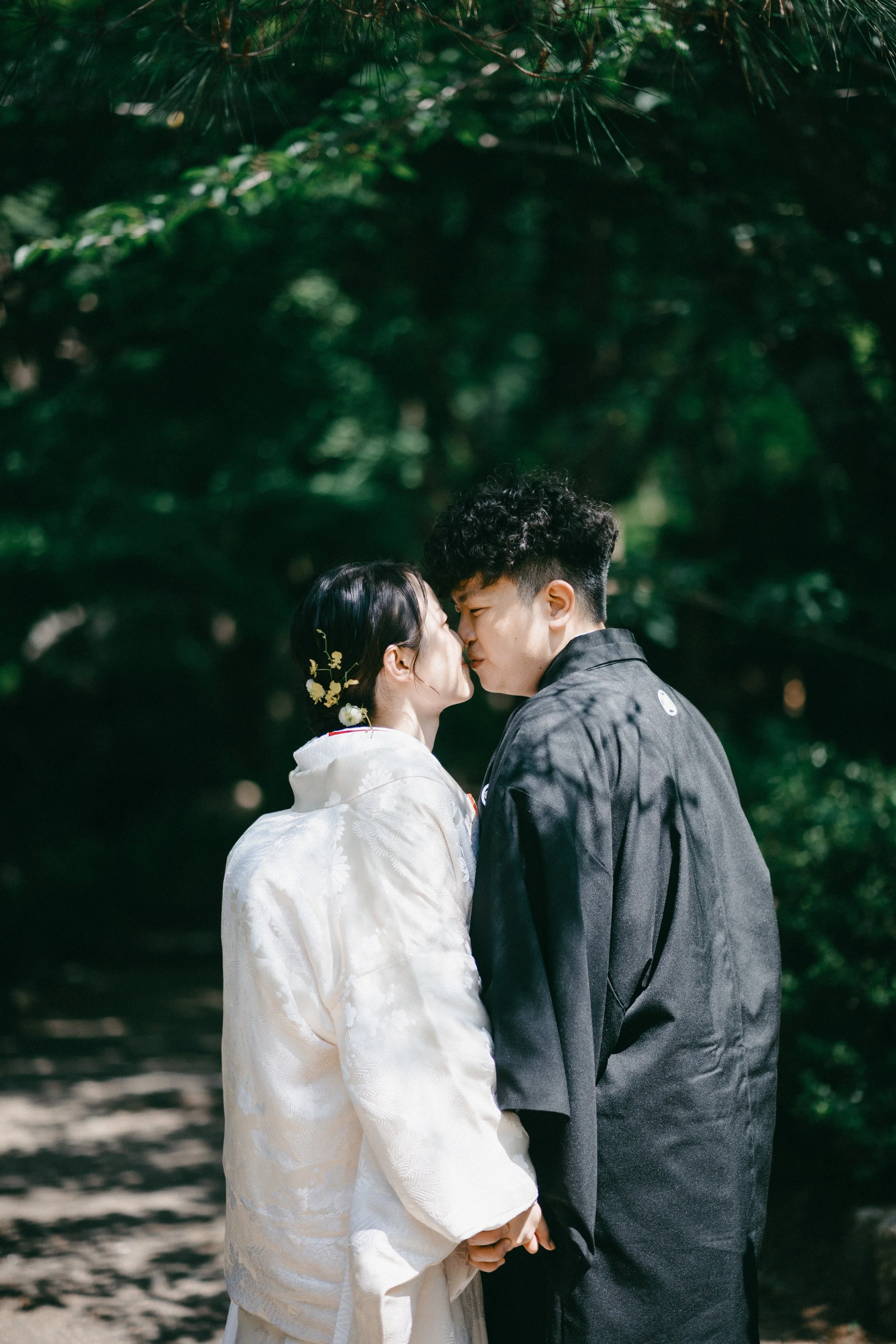 A couple dressed in traditional Japanese wedding attire sharing a kiss outdoors surrounded by green trees.
