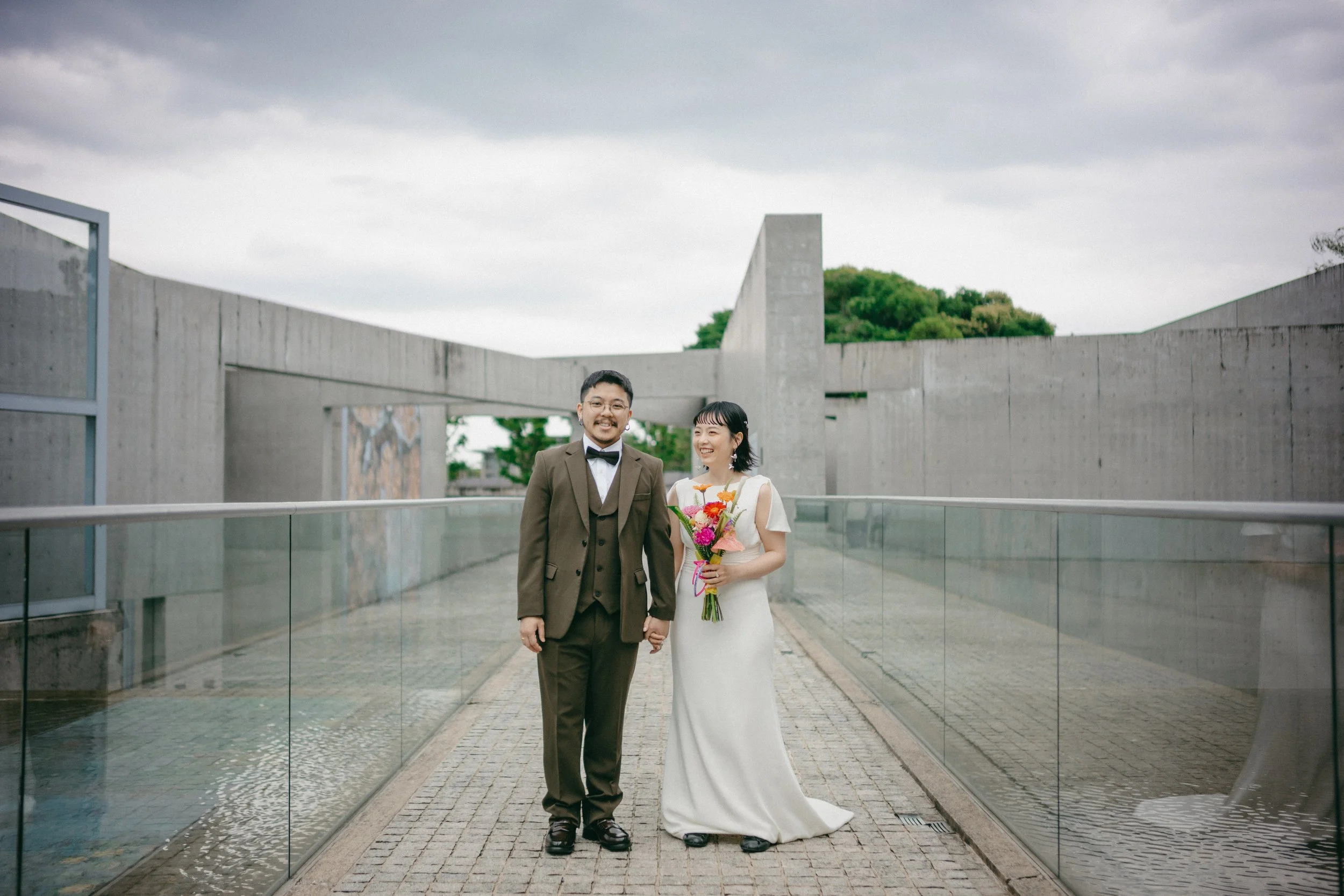 A newlywed couple standing on a bridge outdoors, holding hands. The groom is wearing a green suit with a bow tie, and the bride is in a white wedding dress holding a colorful bouquet. The background features modern concrete structures and greenery un