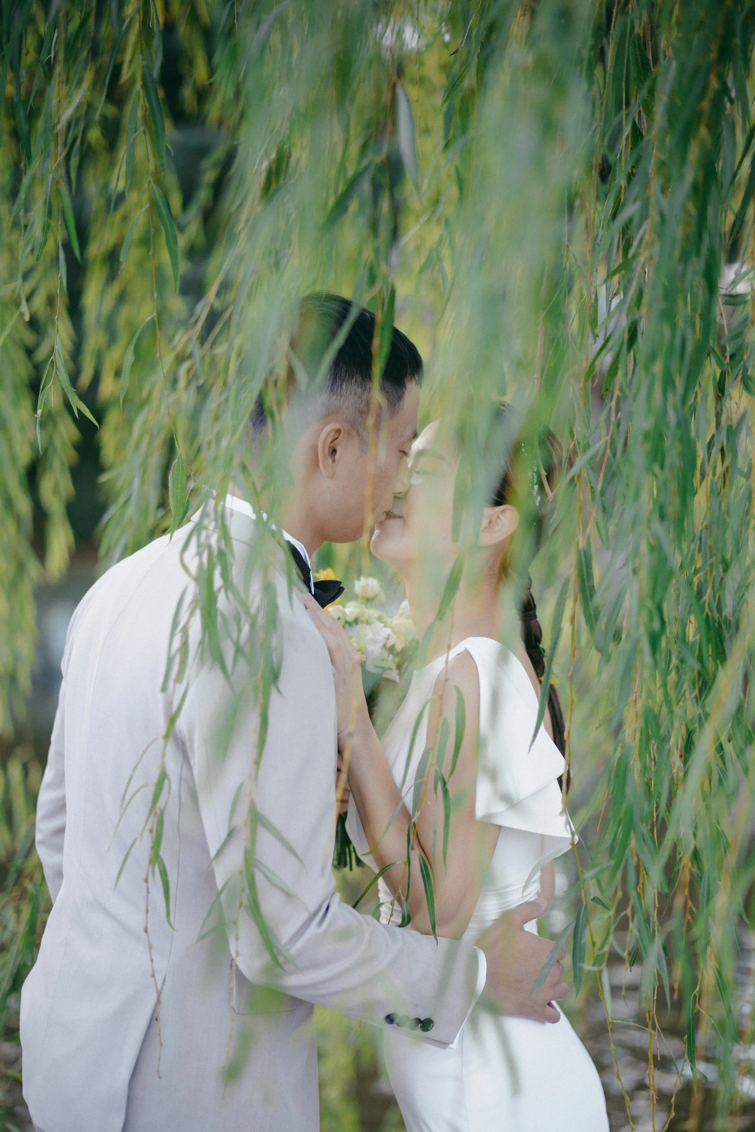 A couple in wedding attire kissing among hanging branches of a tree.