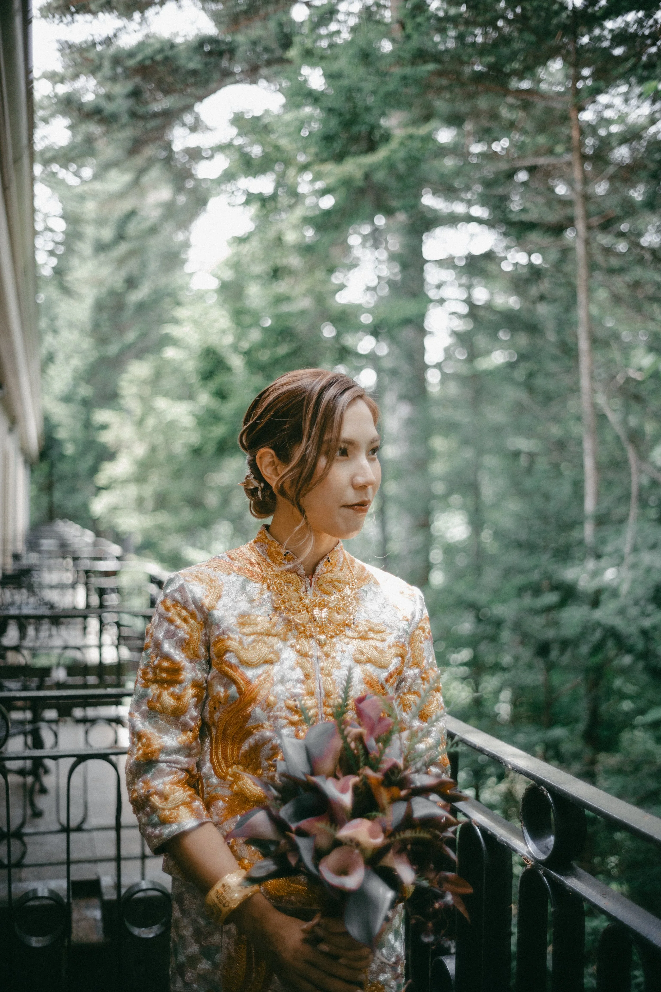 A woman in an ornate traditional dress holding a bouquet of flowers on a balcony overlooking a forested area.