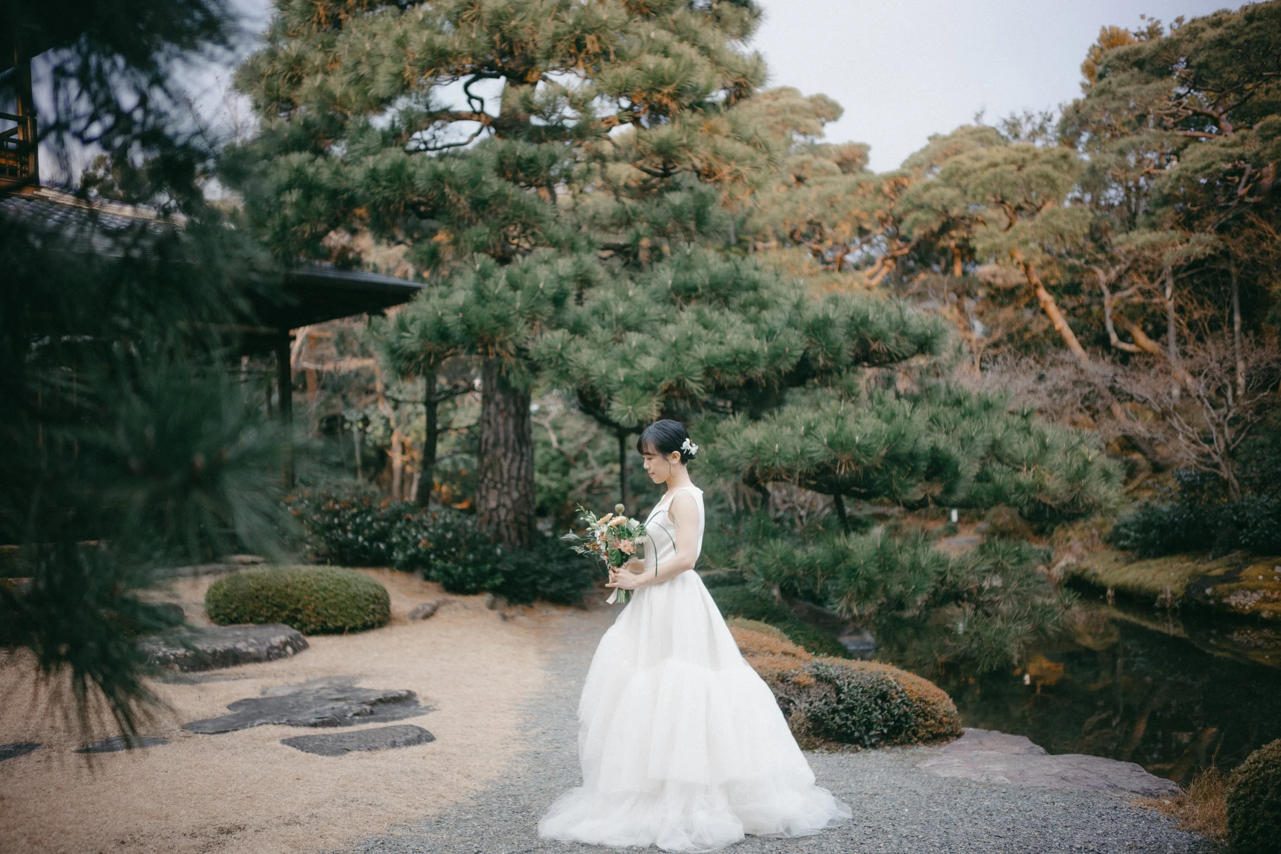 A bride in a white wedding dress holding a bouquet of flowers, standing in a Japanese garden with tall trees and landscaped bushes.