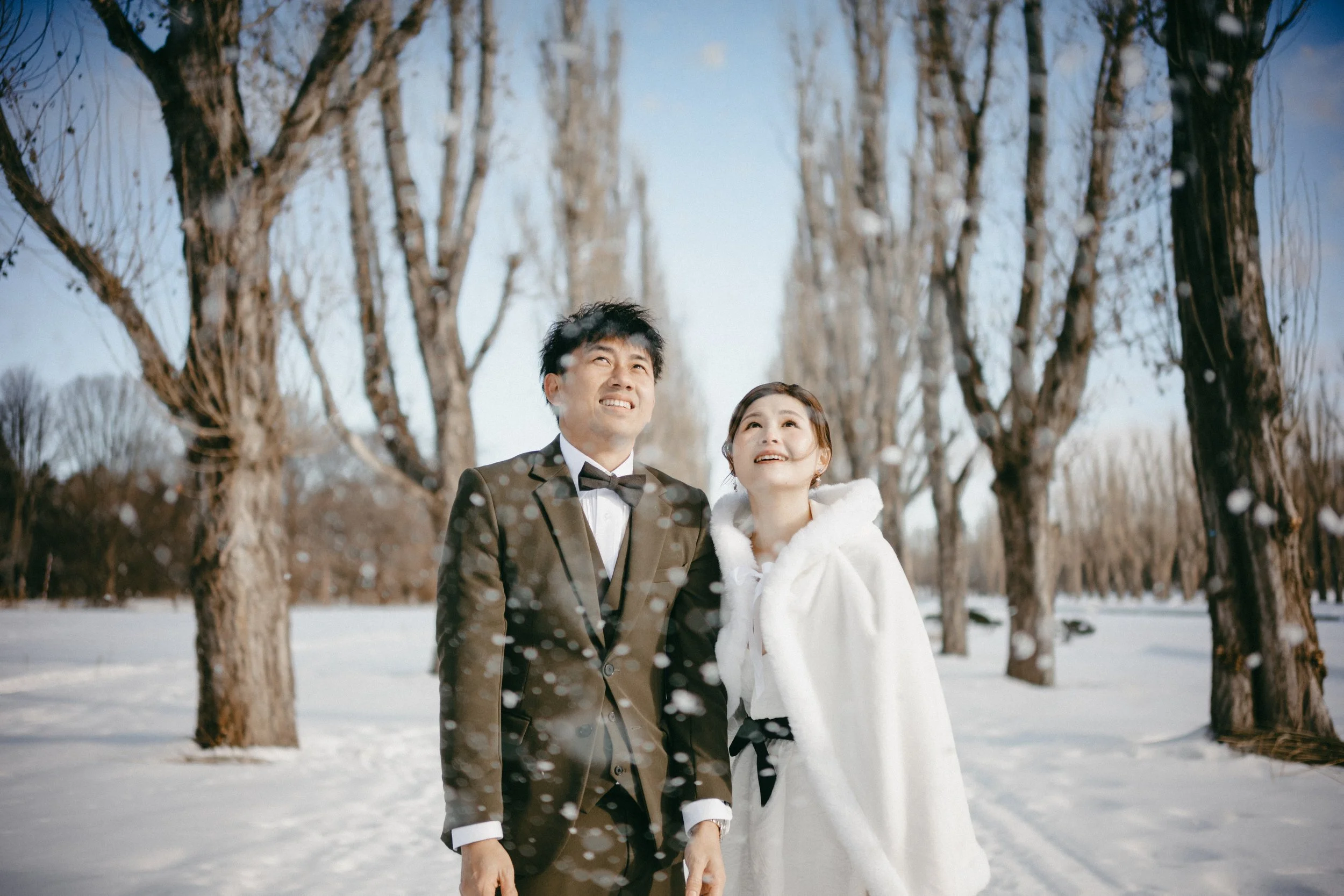 A newlywed couple smiling and looking up while walking in a snowy park surrounded by tall, leafless trees.