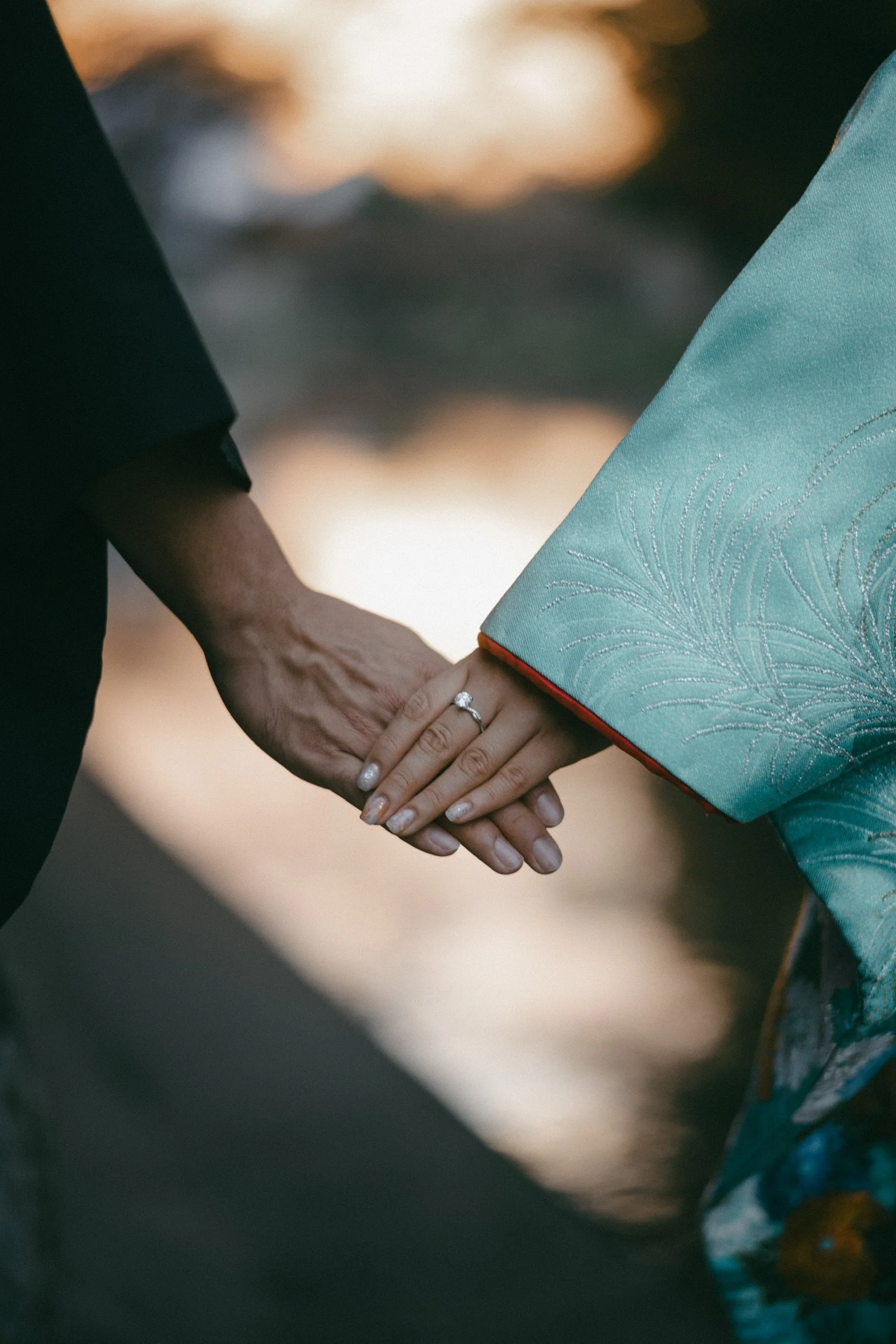 Close-up of a couple holding hands, with a wedding ring visible on the woman's finger, outdoors with a blurred natural background.