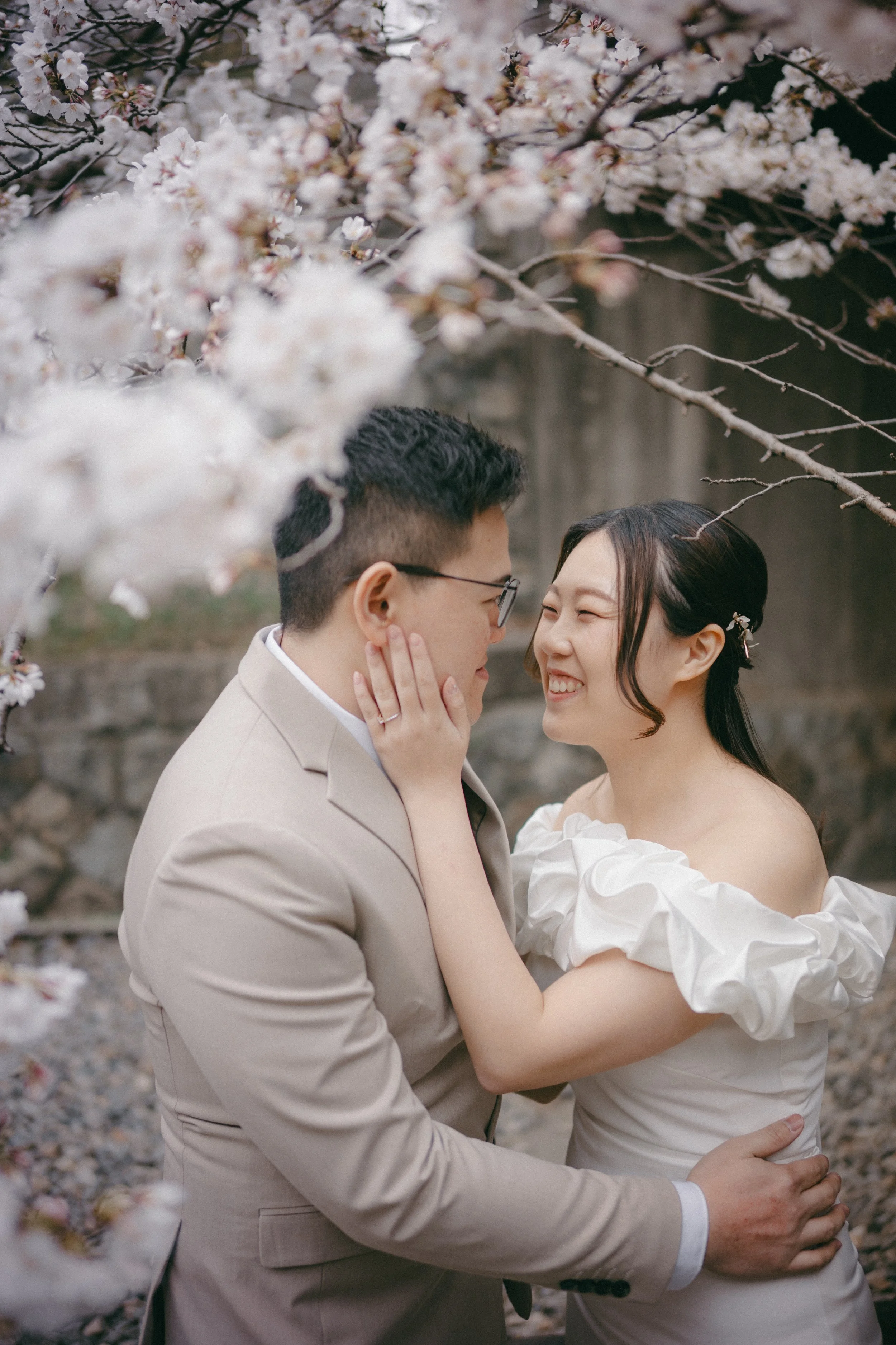 A couple in wedding attire sharing a close, joyful moment under blooming cherry blossom trees.