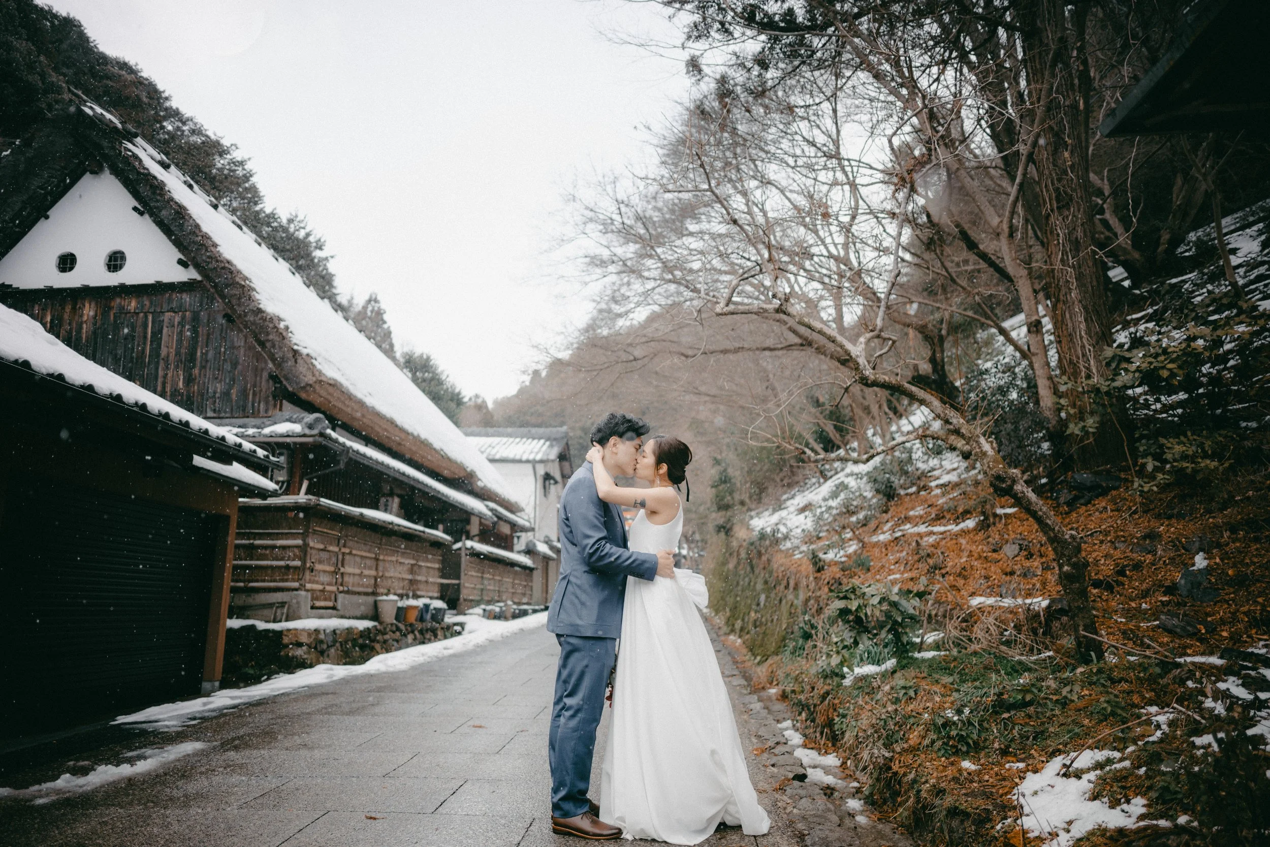 A couple in wedding attire kissing on a snowy street with traditional wooden houses and leafless trees in the background.