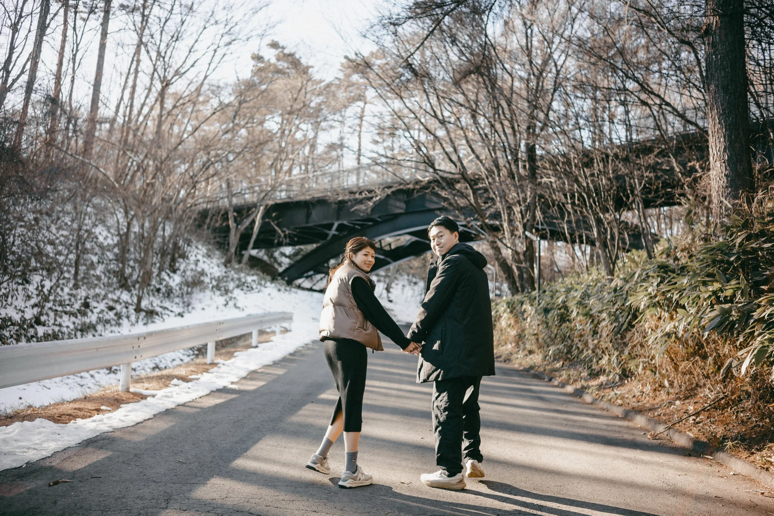 A couple holding hands and smiling on a snow-dusted path in a wooded area during winter.