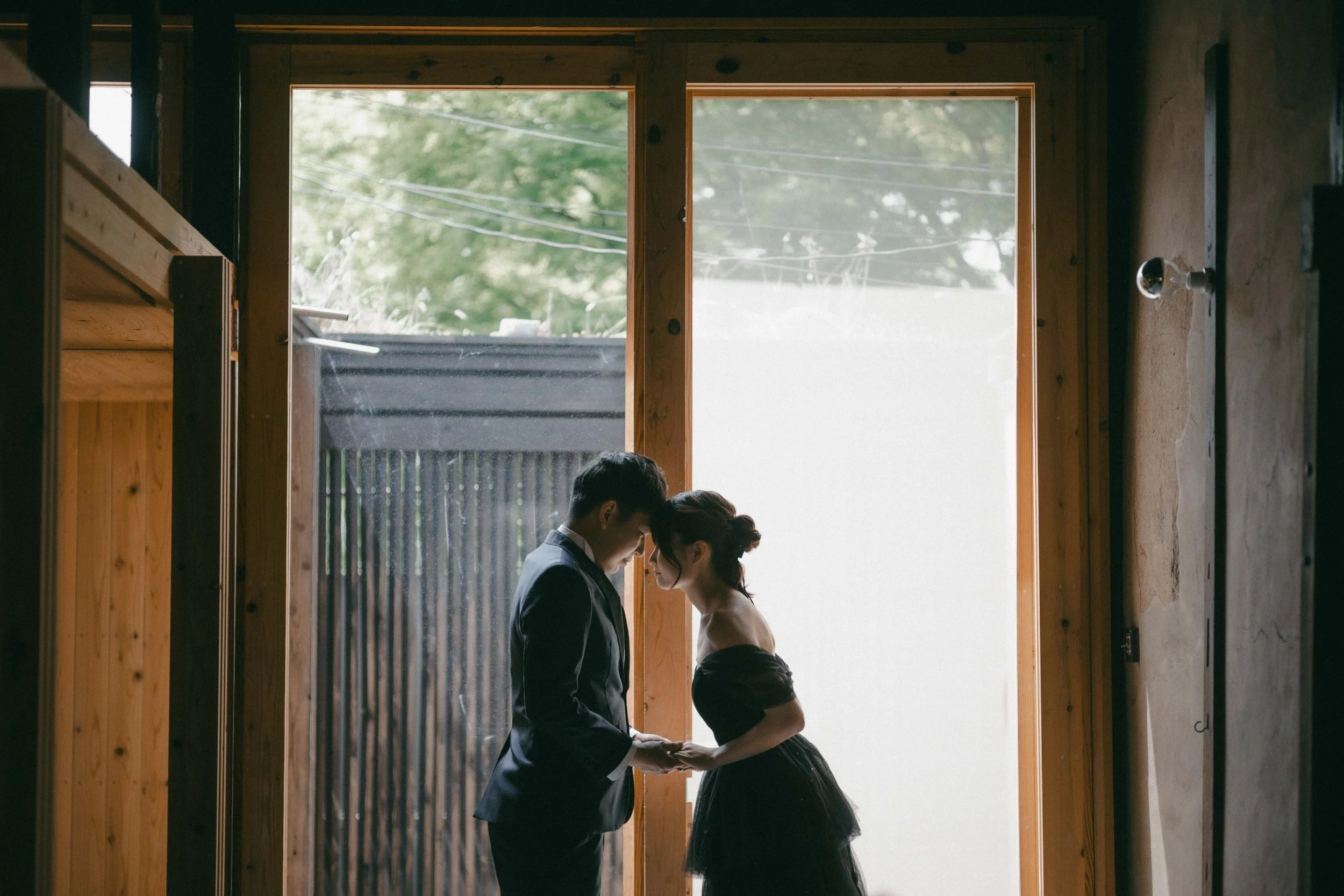 A romantic moment between a man and woman dressed for a special occasion, holding hands and touching foreheads inside a rustic wooden room with large windows and outdoor greenery.