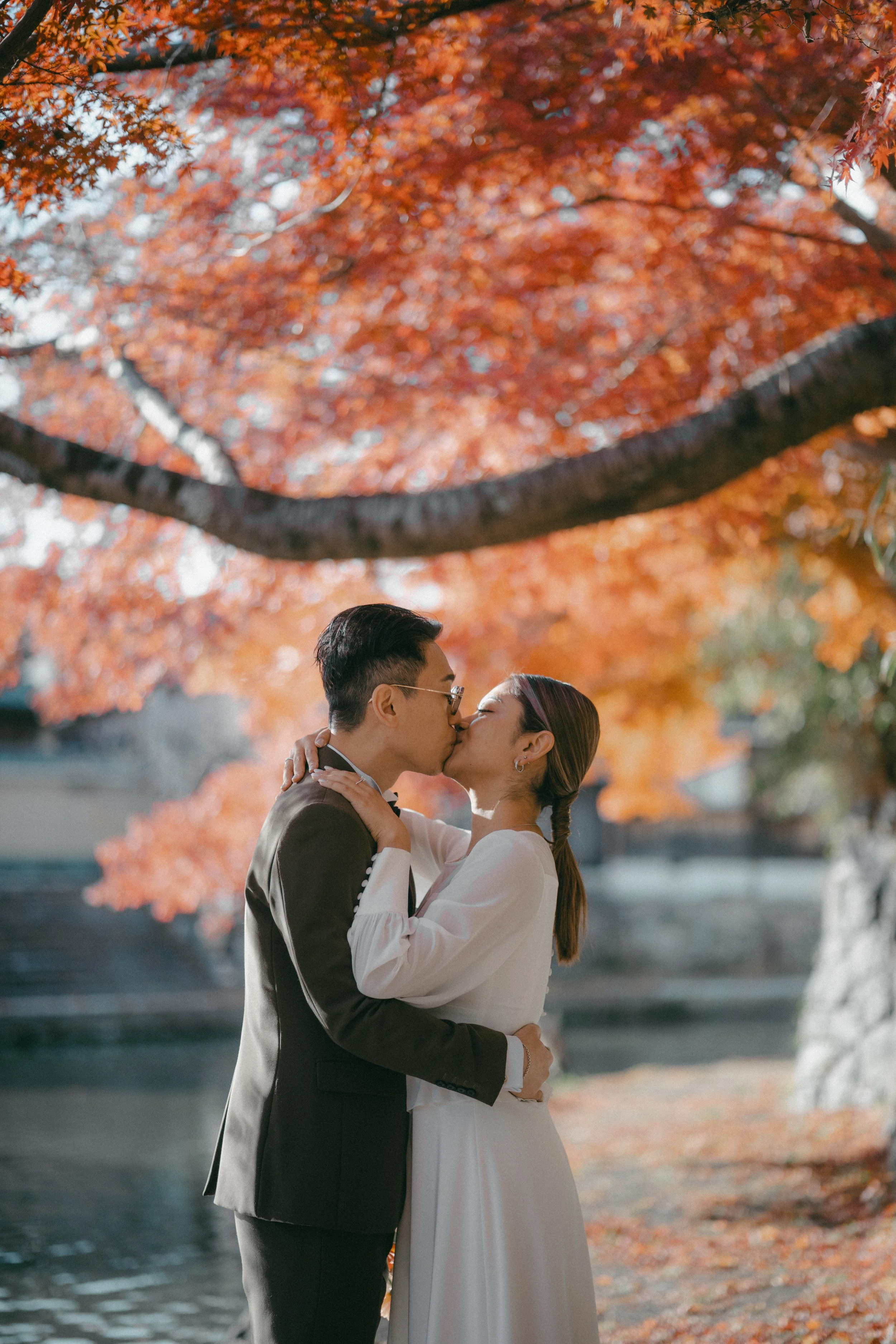 A couple kissing in front of autumn-colored trees and a pond.