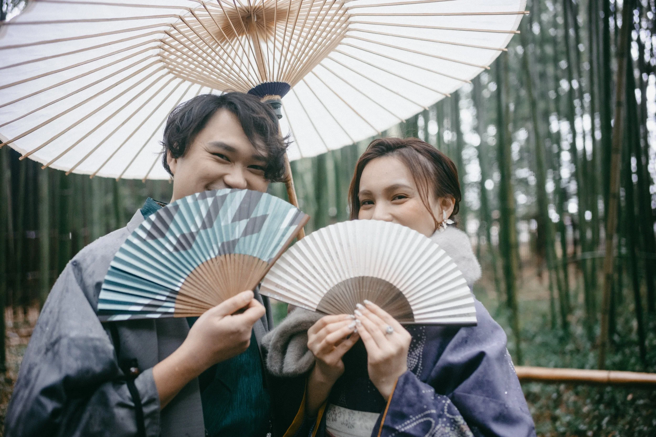 Two young people standing in a bamboo forest holding traditional handheld fans and a parasol, smiling and pretending tohide their faces behind the fans.