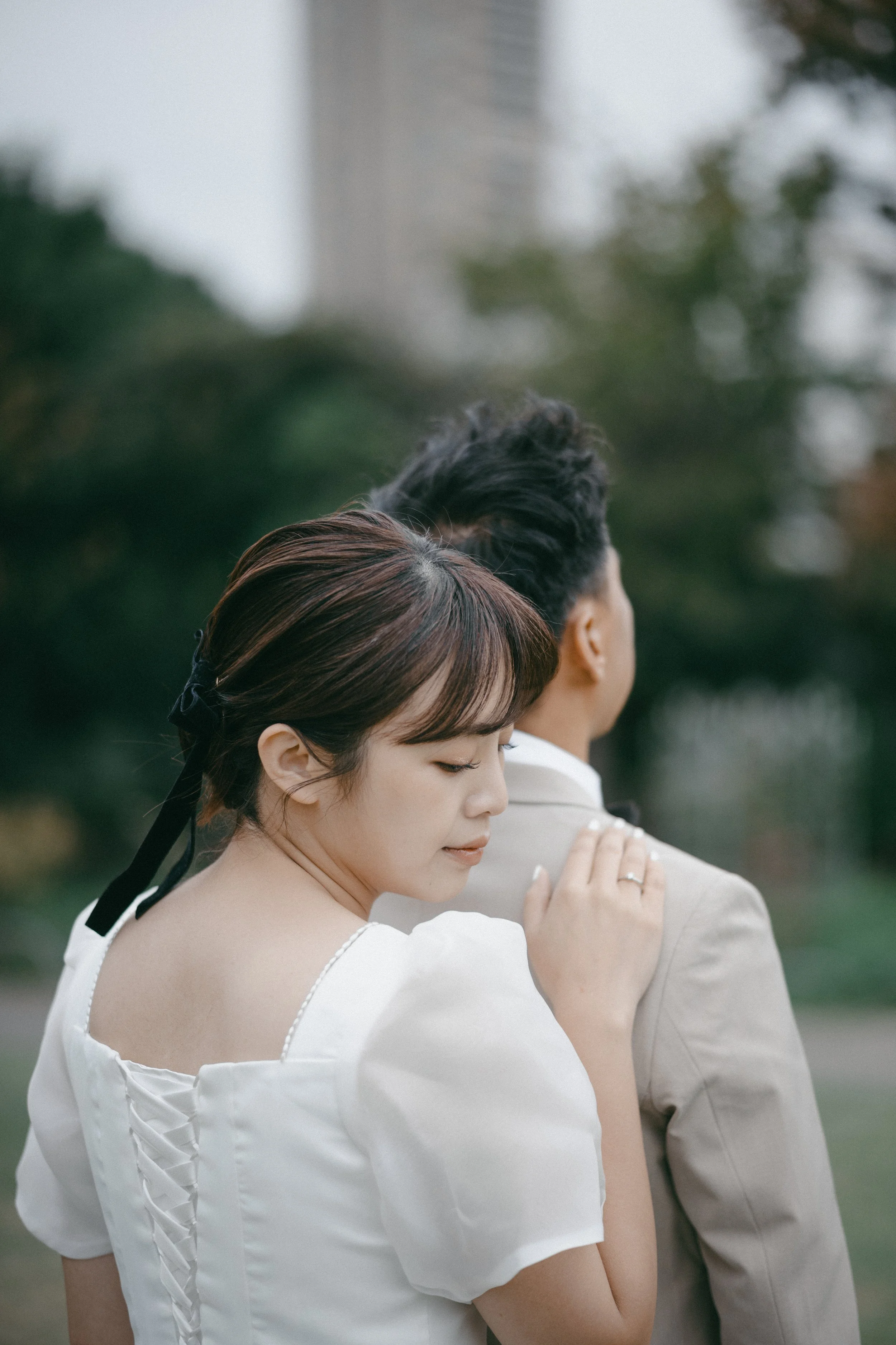 A woman resting her head on the shoulder of a man, both standing outdoors, with trees and a blurred building in the background. The woman is wearing a white dress with puffed sleeves and has a black ribbon in her hair.
