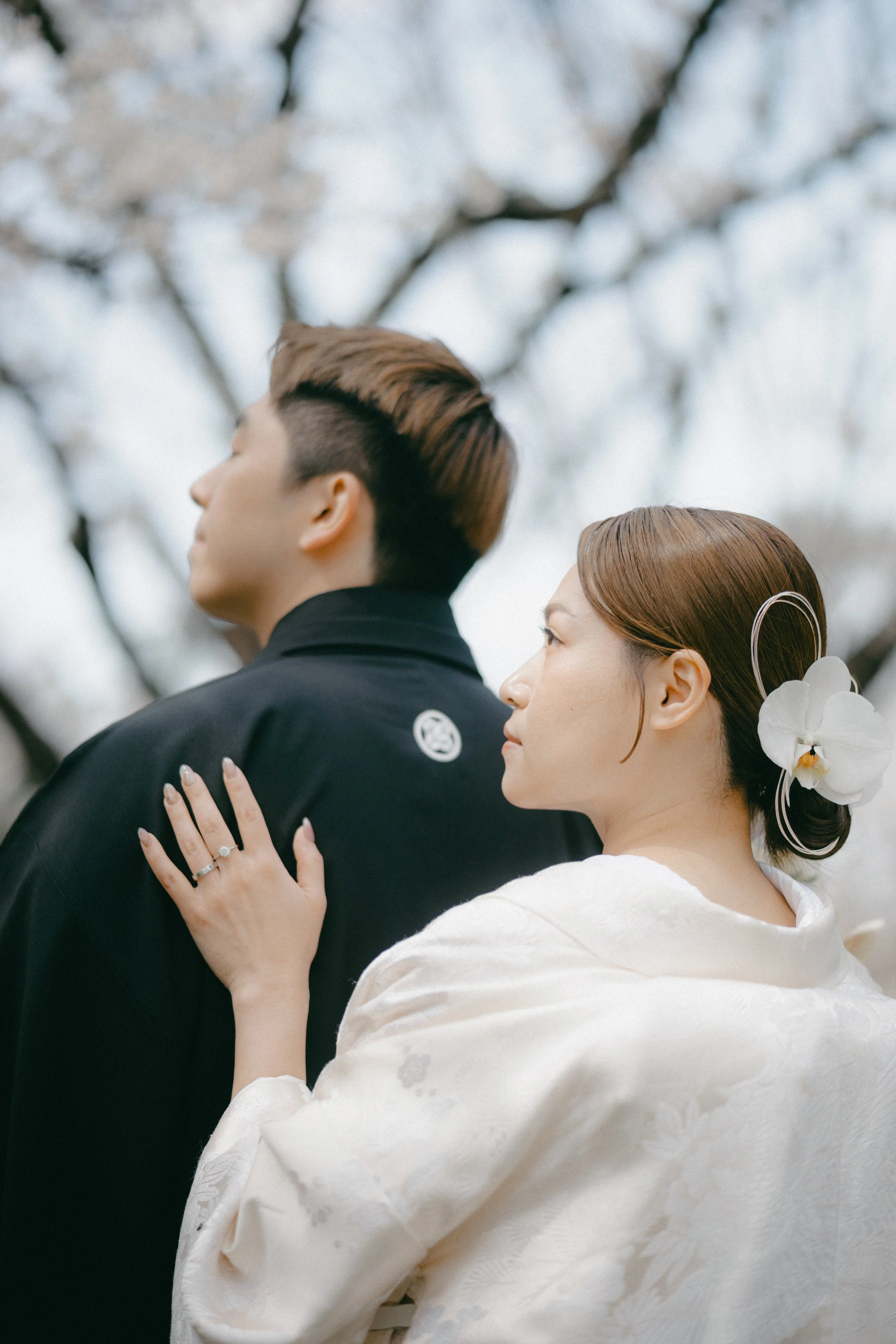 A couple dressed in traditional Japanese wedding attire, with the woman wearing a white kimono and floral hair accessory, and the man in a black kimono, standing outdoors with cherry blossoms in the background.