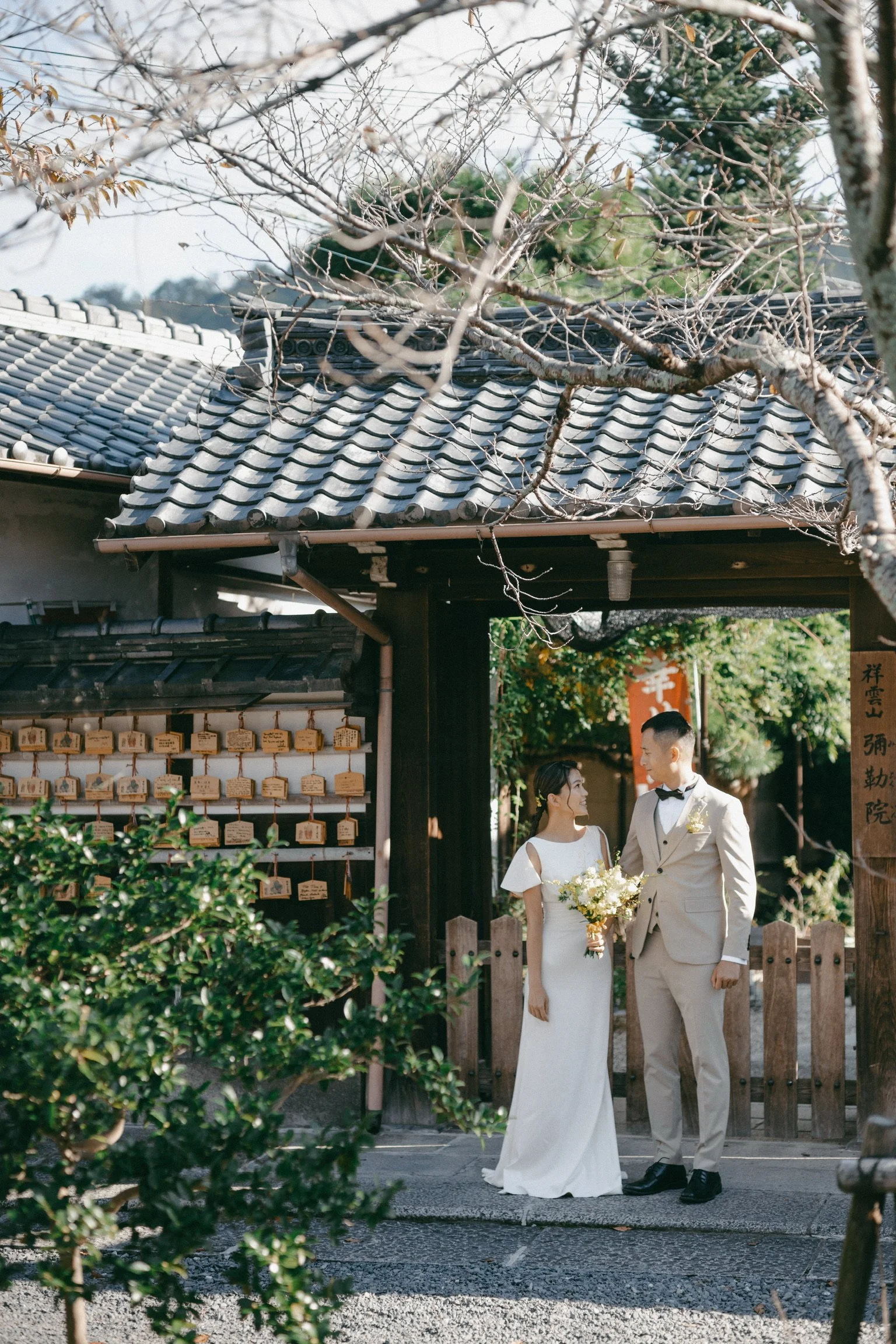 A newlywed couple standing outside a traditional Japanese building, the bride holding a bouquet and gazing at the groom, surrounded by greenery and wooden signs.