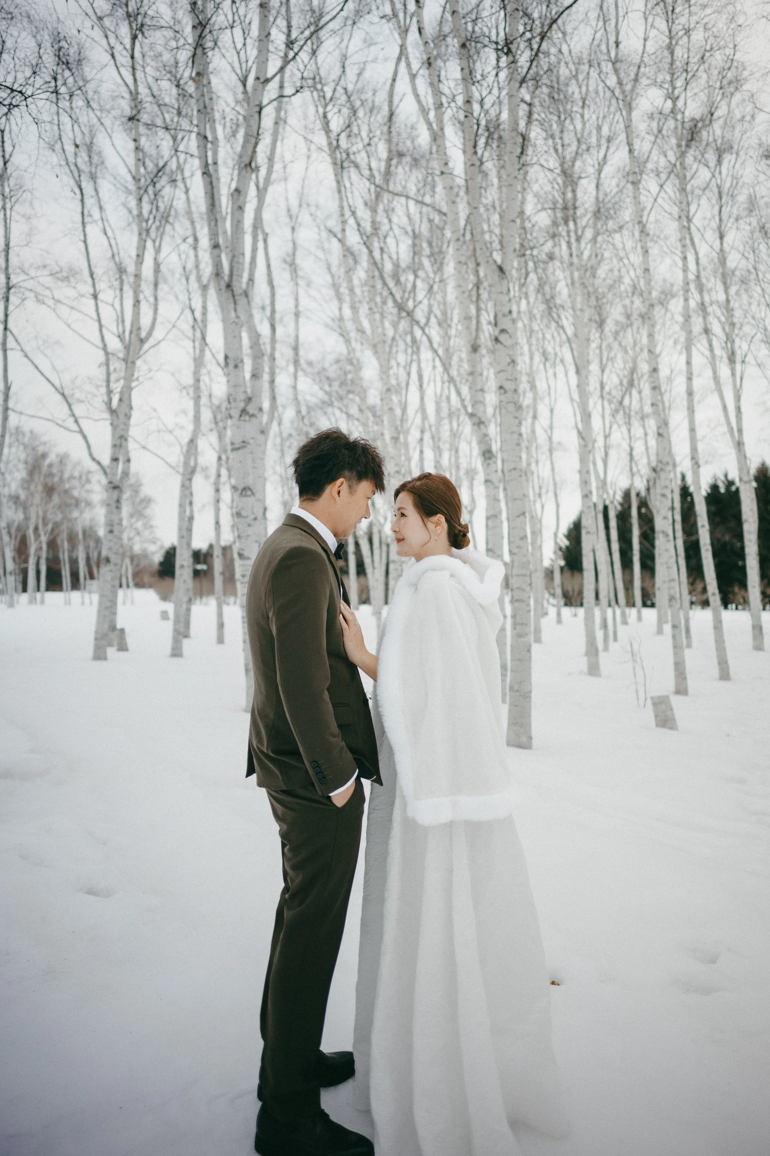 A couple in wedding attire standing face to face in a snow-covered forest with bare trees.