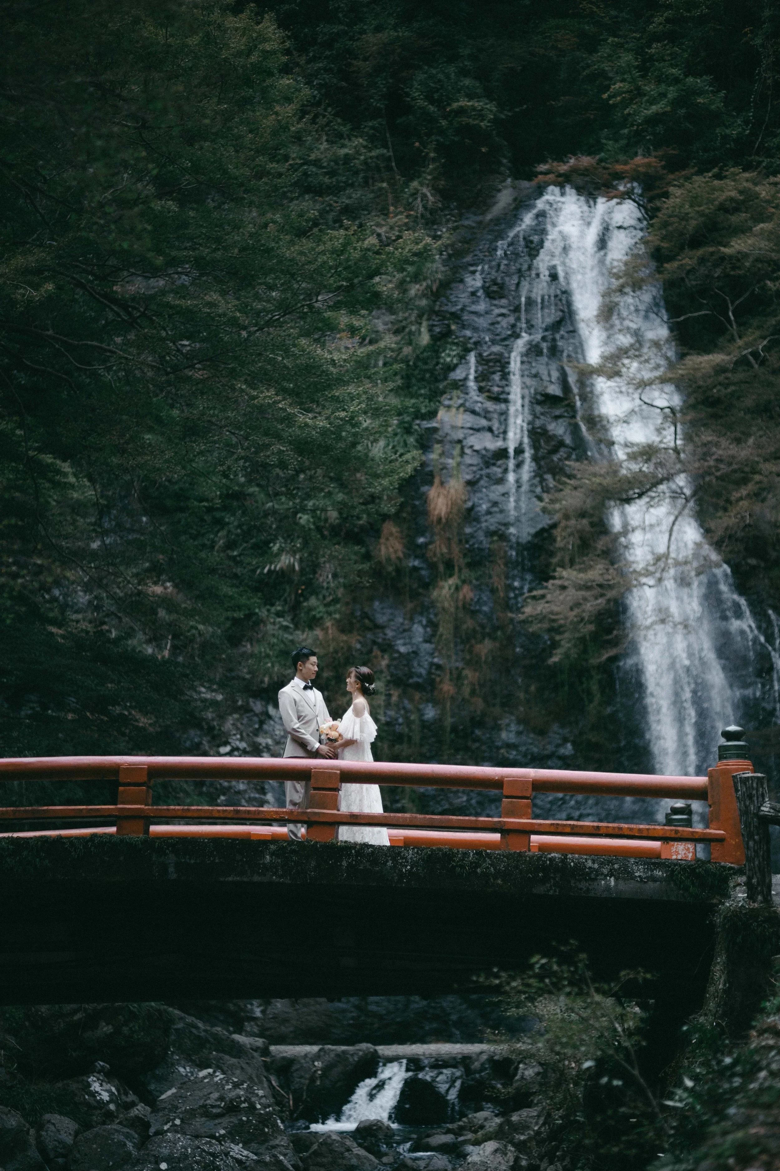 A bride and groom standing on a bridge in front of a waterfall surrounded by lush greenery.