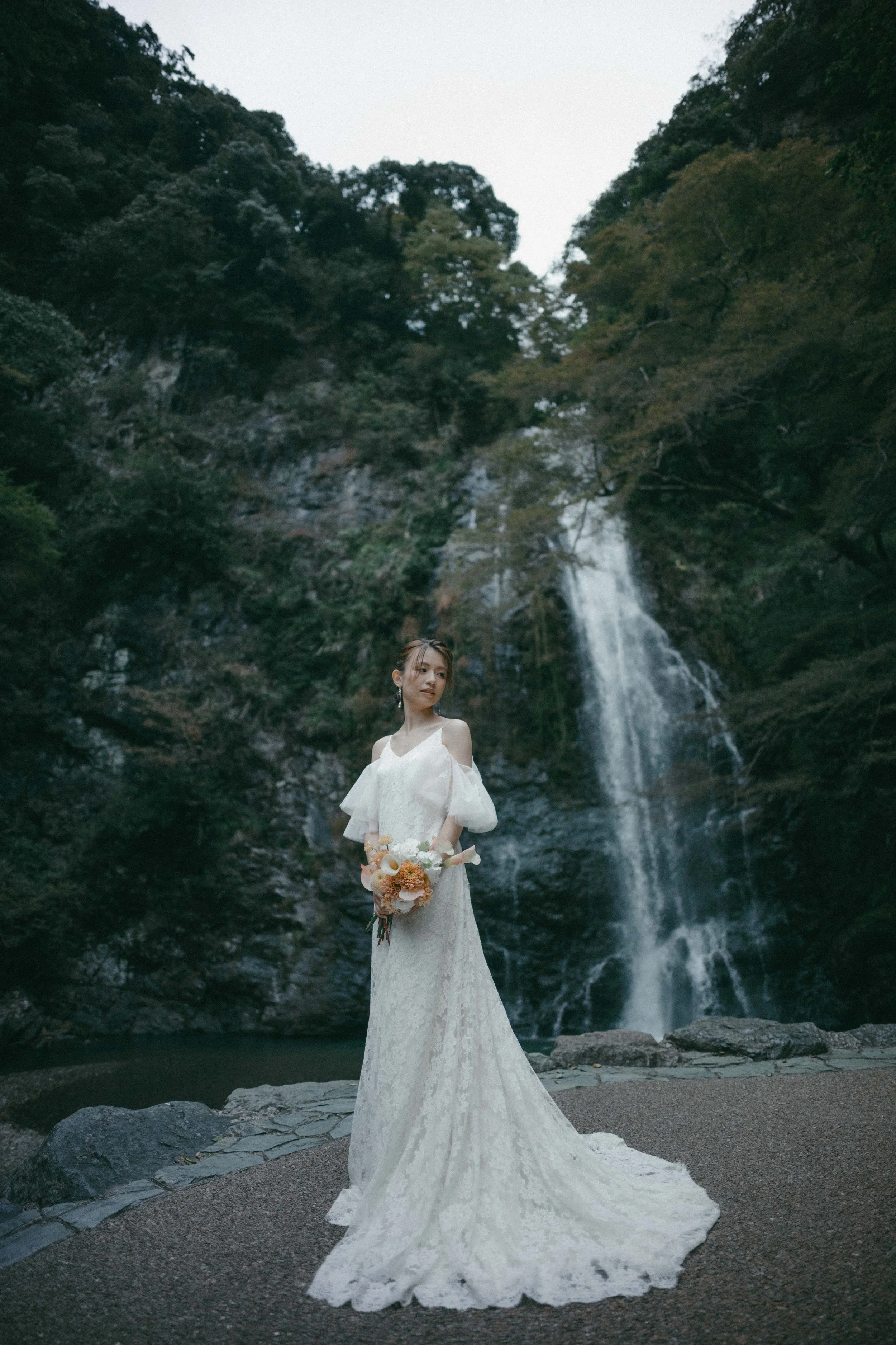 A woman in a white wedding dress holding a bouquet of flowers, standing outdoors near a waterfall and lush green trees.