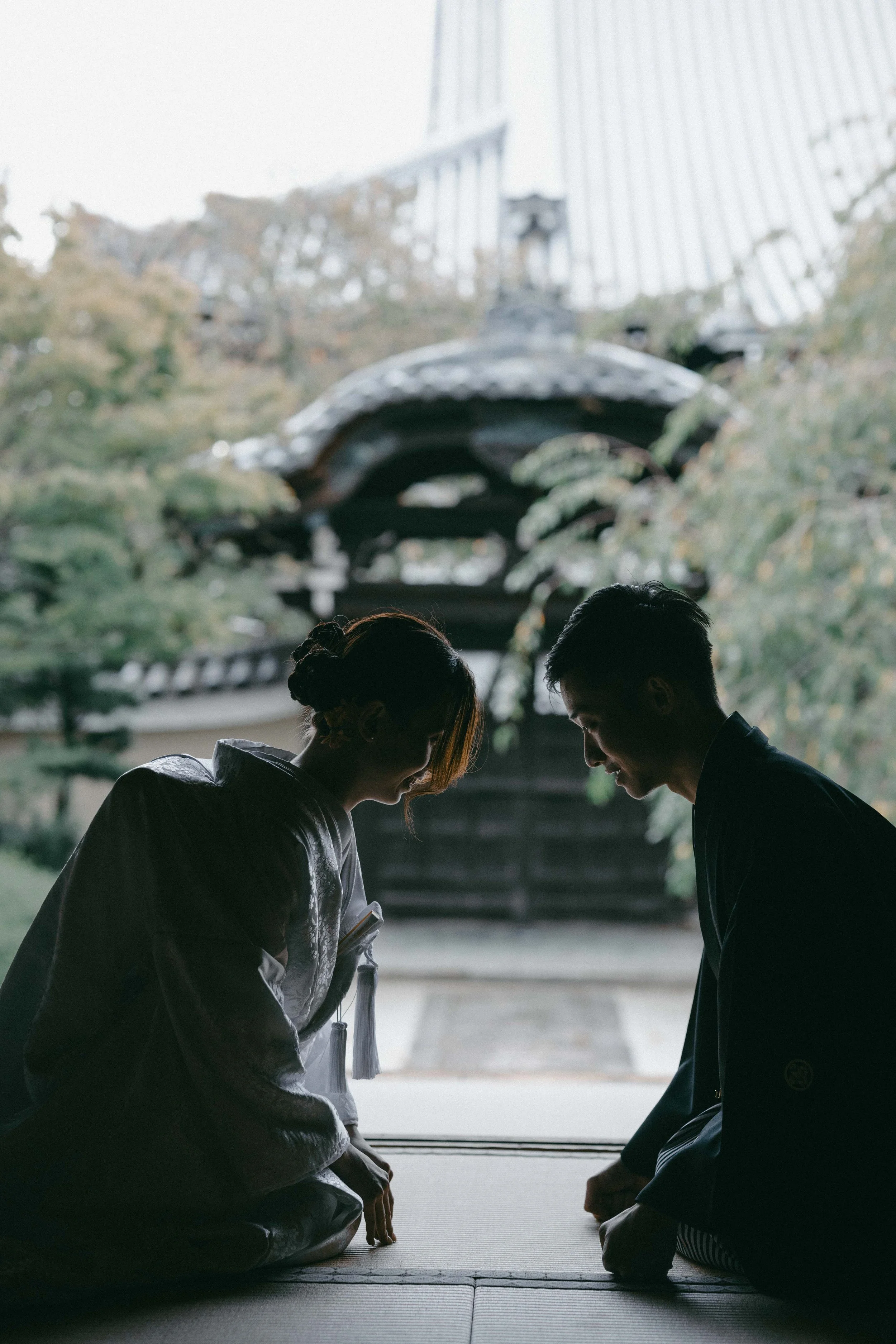 Silhouettes of a man and woman in traditional Japanese clothing bowing to each other inside a temple, with a traditional roof and trees visible in the background.