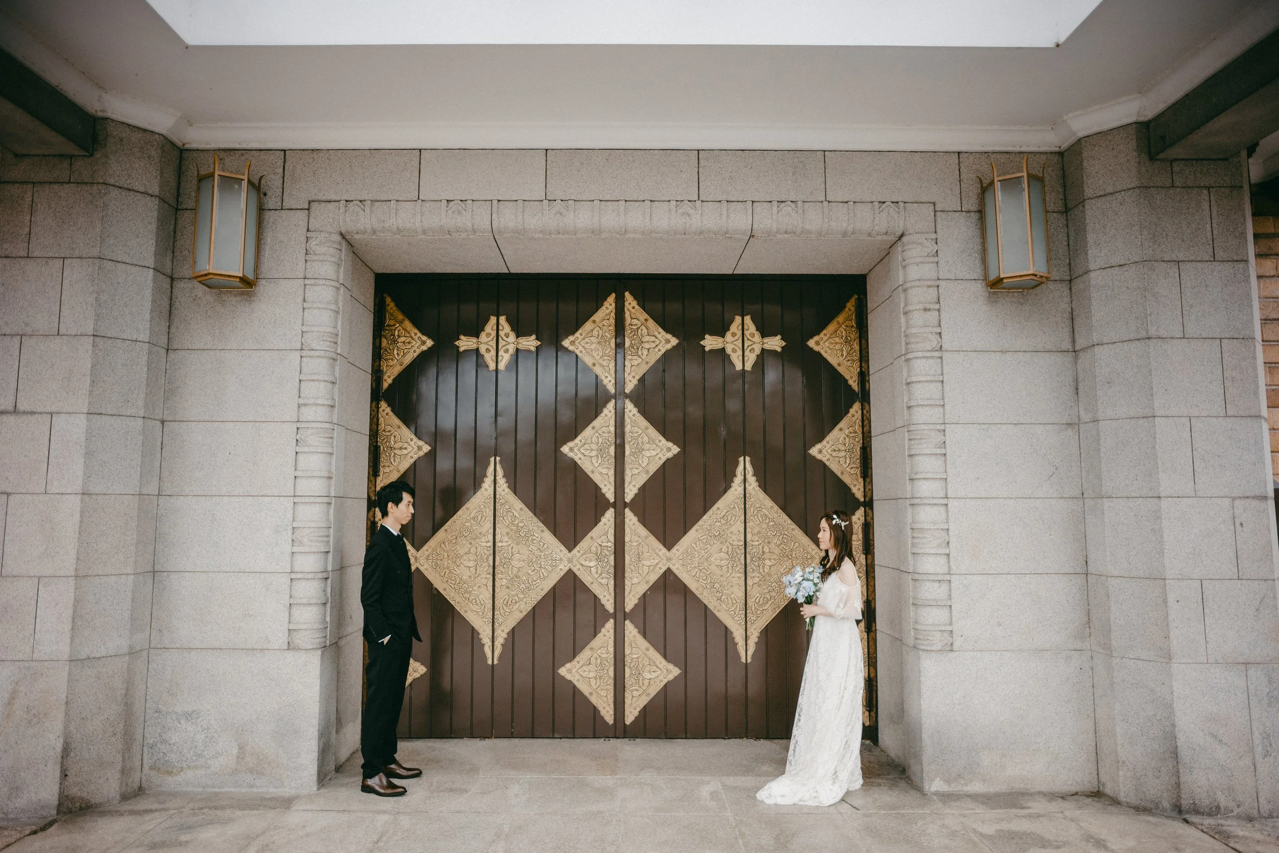 A bride in a white wedding gown holding a bouquet of flowers and a groom in a black suit standing apart in front of large decorative doors with gold accents, outside a stone building.