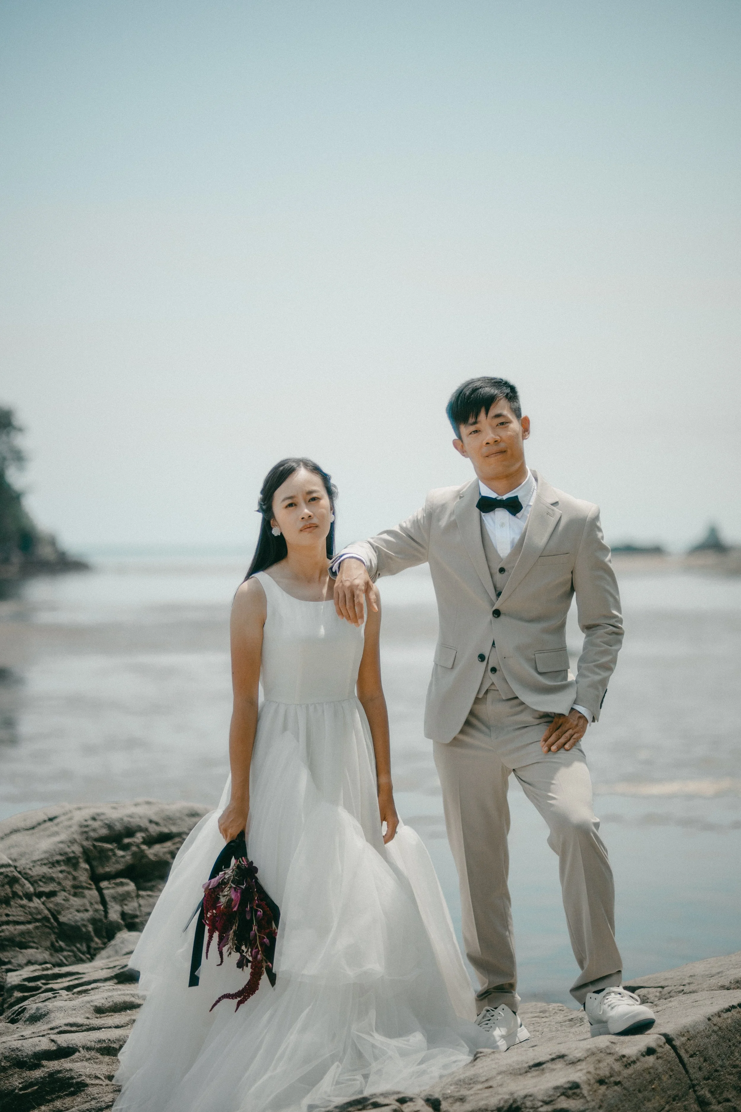 A bride and groom stand on rocks by the beach, with the bride in a white wedding dress holding a bouquet and the groom in a beige suit with a black bow tie, the groom leaning on the bride's shoulder.