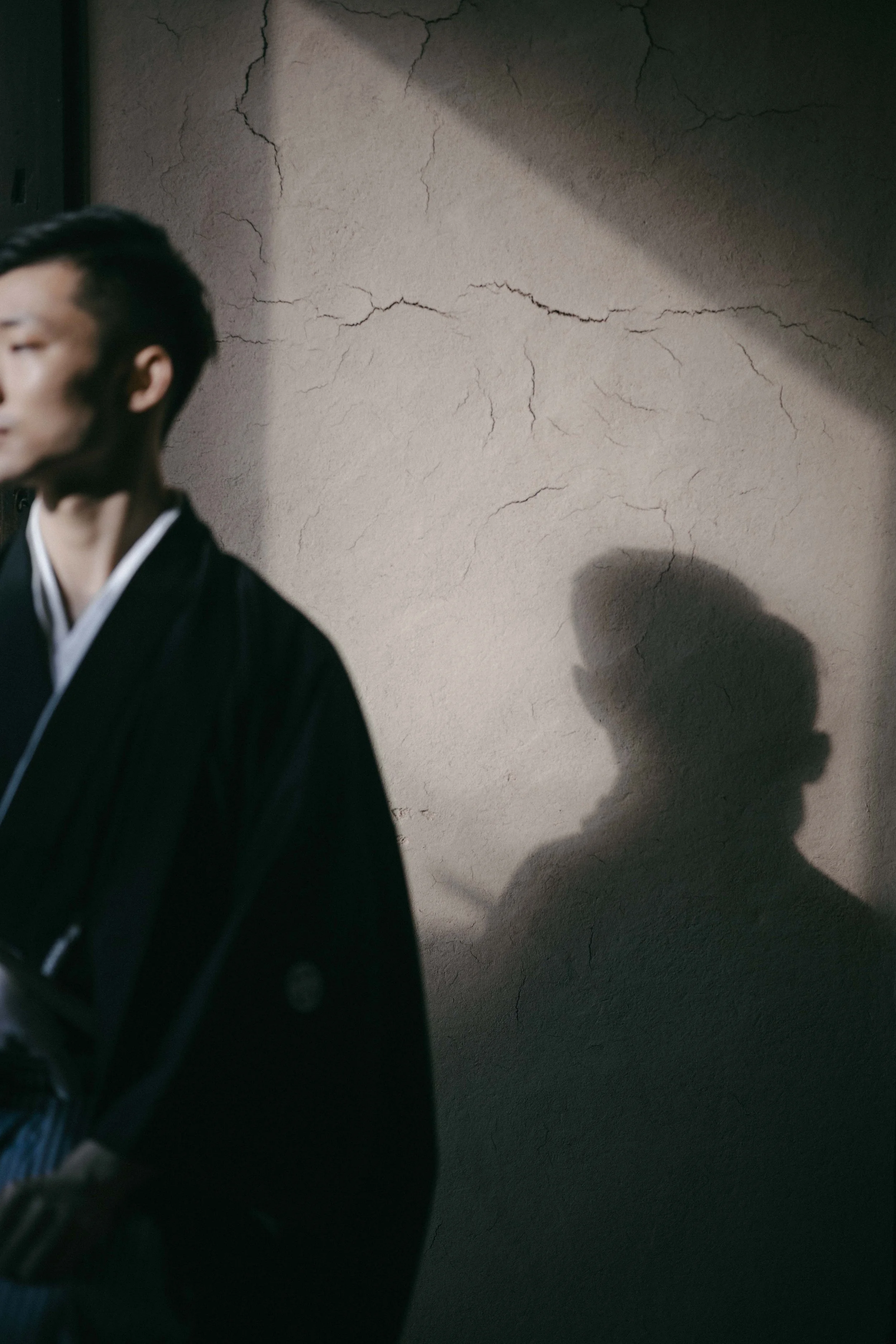 A man in traditional Japanese attire standing in front of a textured wall with prominent cracks, with his shadow cast on the wall behind him.