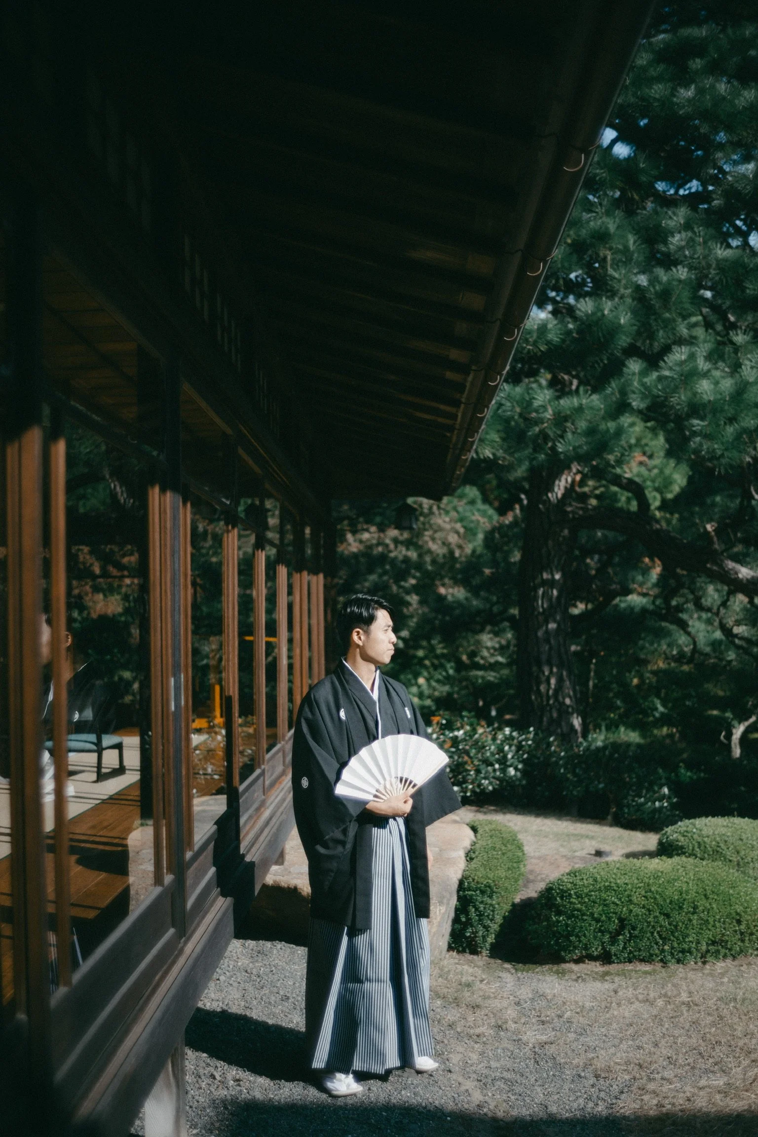 A man dressed in traditional Japanese attire stands outdoors holding a fan, next to a wooden building with large windows and surrounded by greenery.