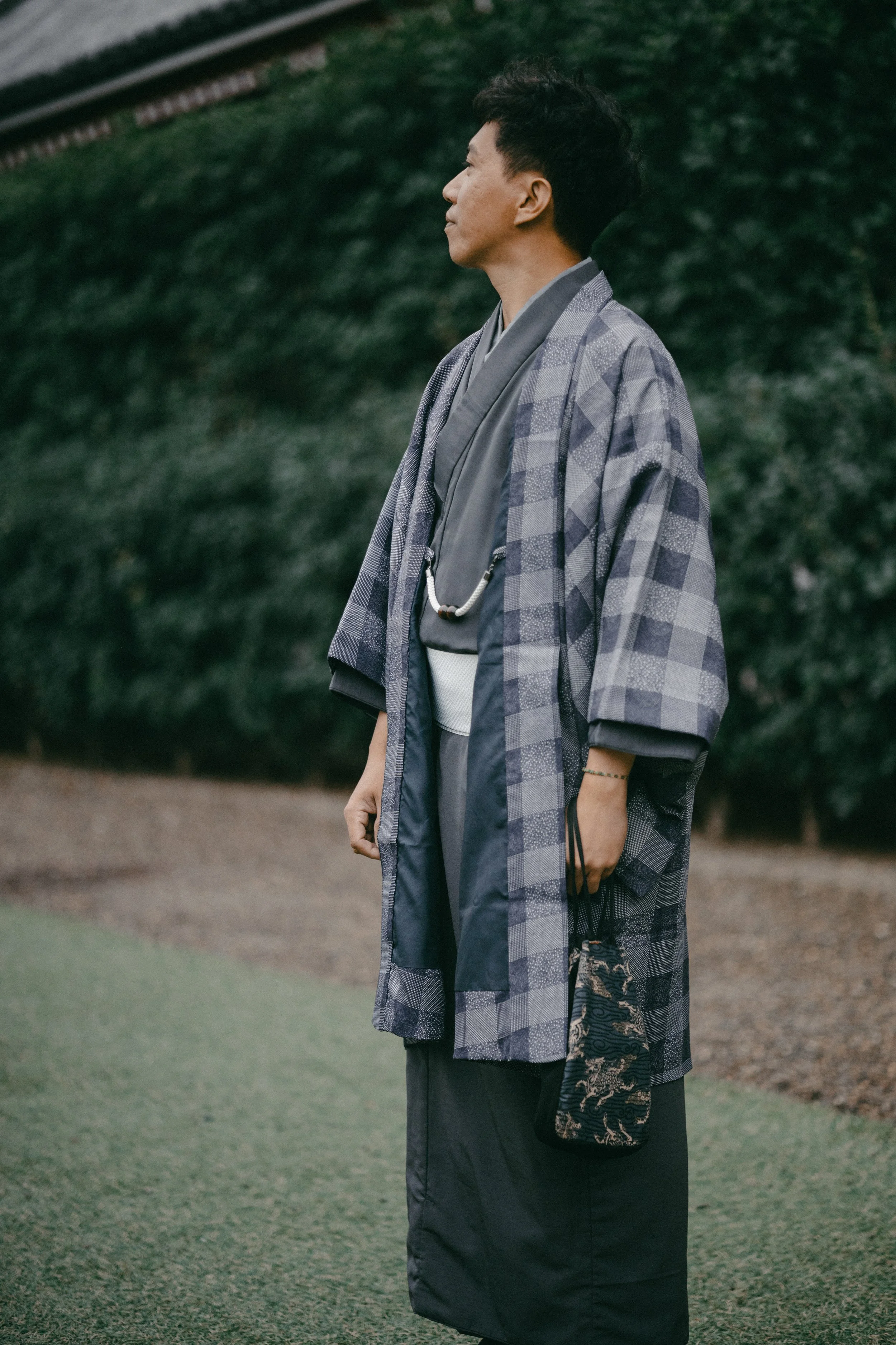 A woman dressed in traditional Japanese clothing, standing outdoors with a background of green trees and bushes.
