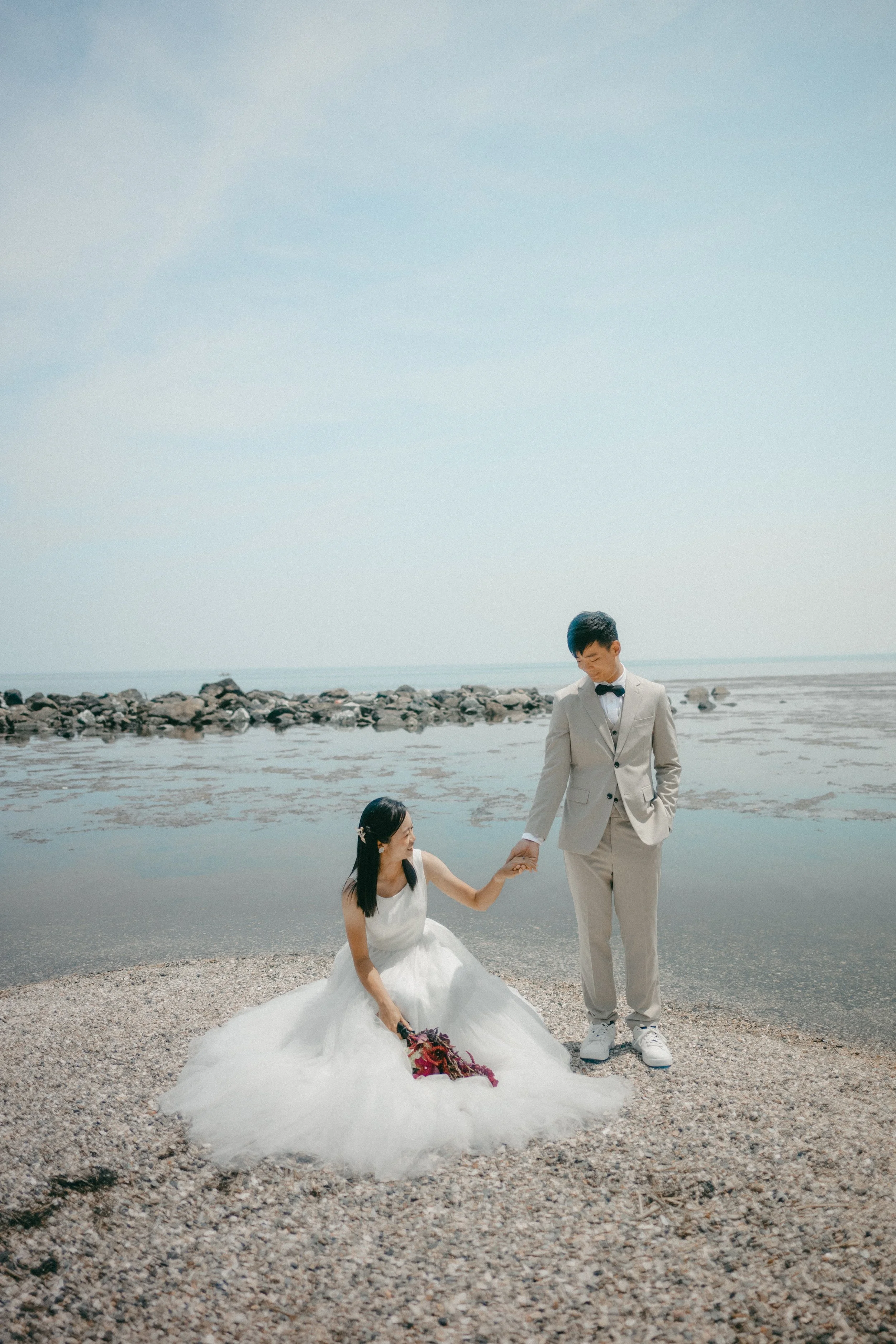 A bride and groom on a beach, with the bride sitting on the sand holding a bouquet, and the groom standing, holding her hand, both dressed in wedding attire.