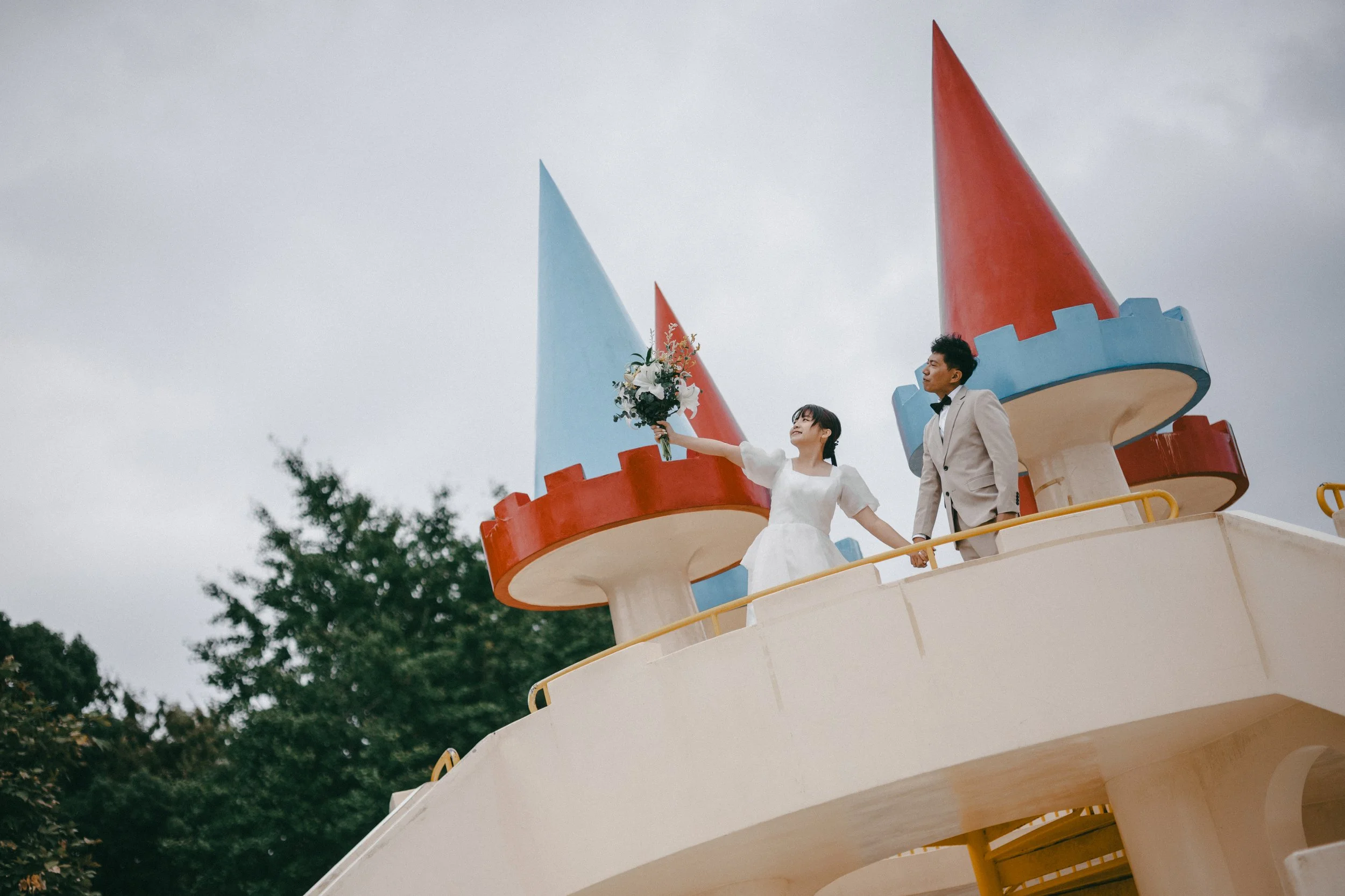 A bride and groom standing on a castle-themed structure at an amusement park, with the bride holding a bouquet and extending it outward, both dressed in wedding attire.