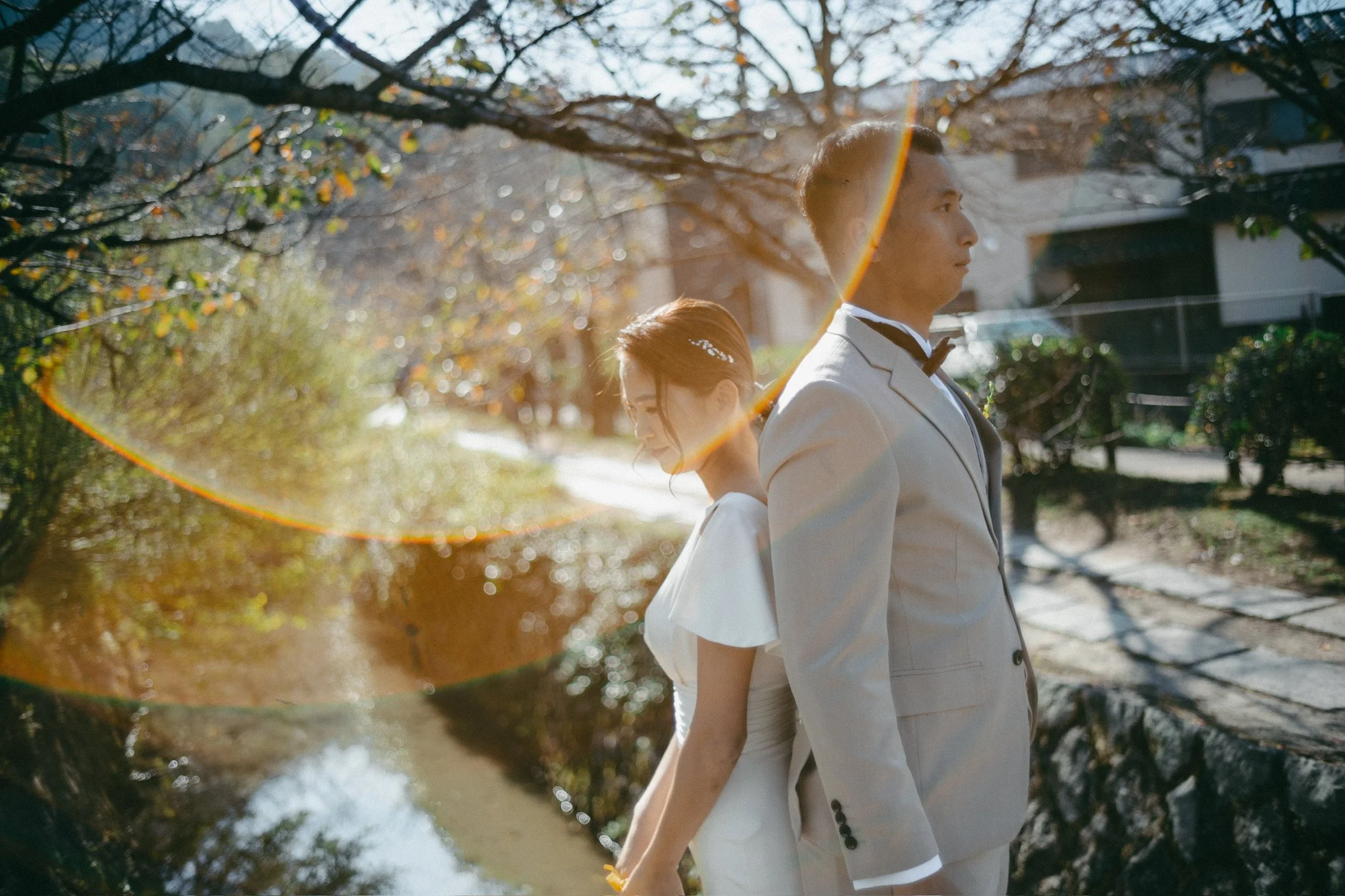A man and woman dressed in formal wedding attire standing back to back outdoors near a stream with trees and buildings in the background, sunny day.