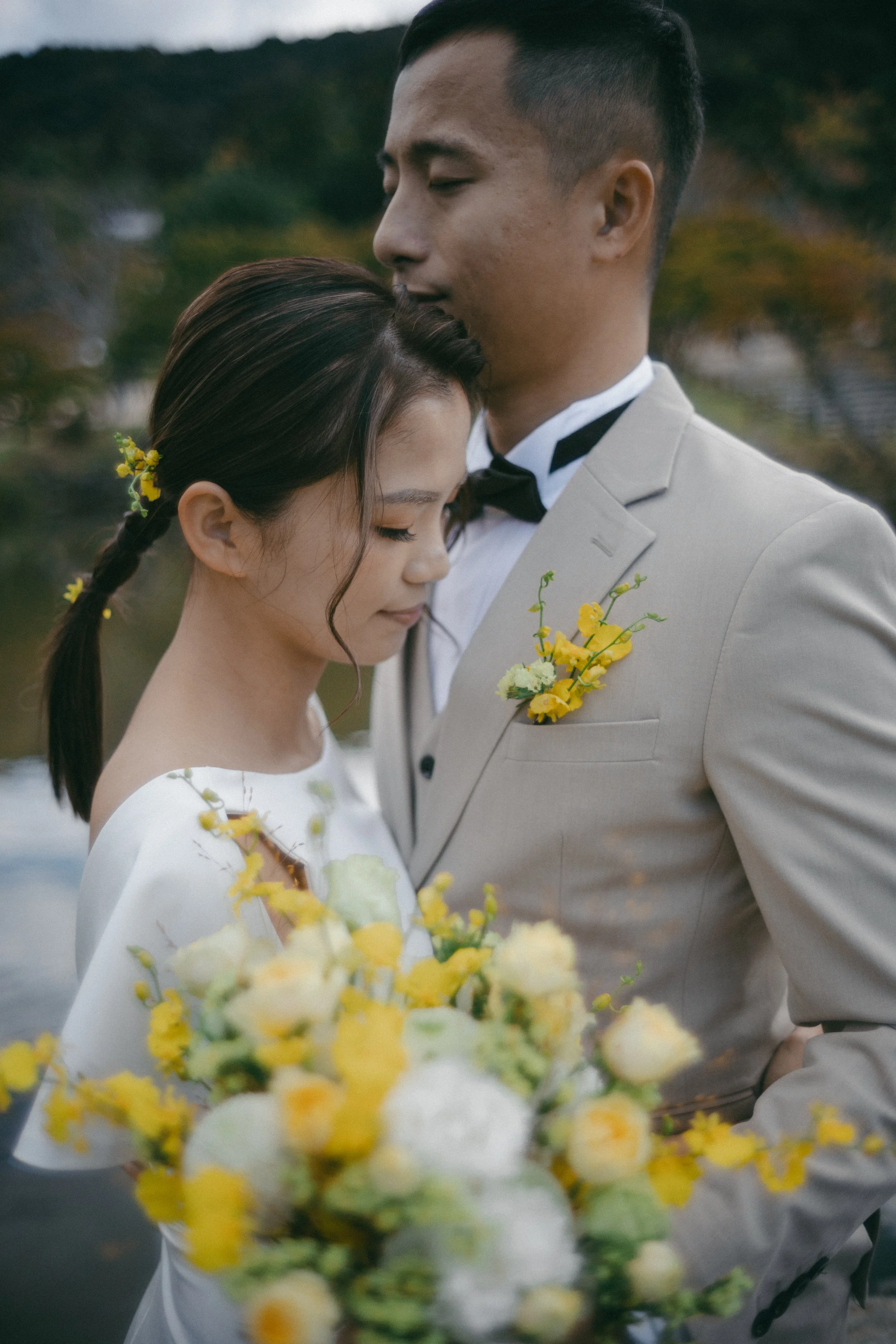 A bride and groom embrace outdoors, with the bride holding a yellow and white floral bouquet, both dressed elegantly.