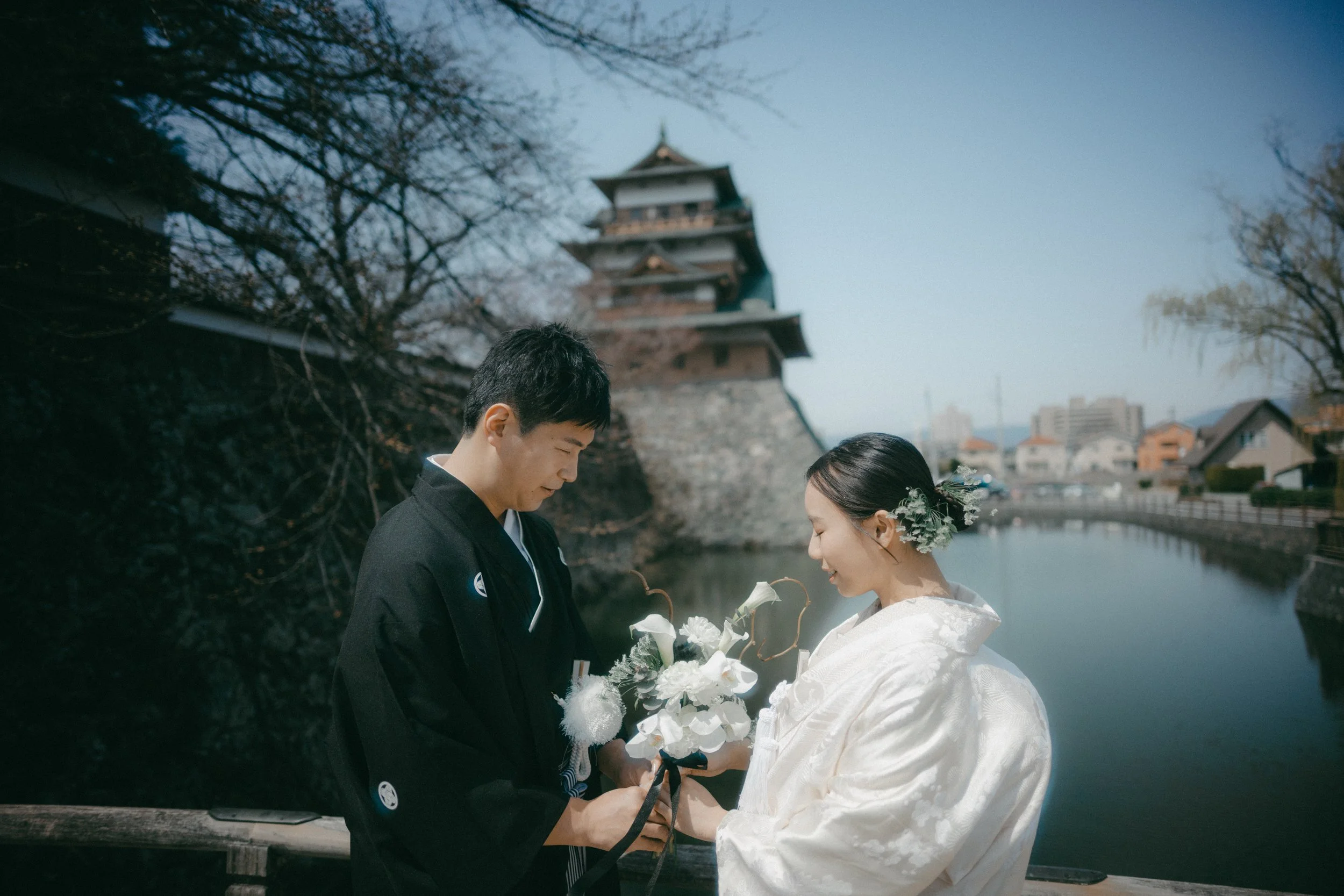 A couple in traditional Japanese wedding attire standing on a bridge by a river, with a historic Japanese castle in the background.