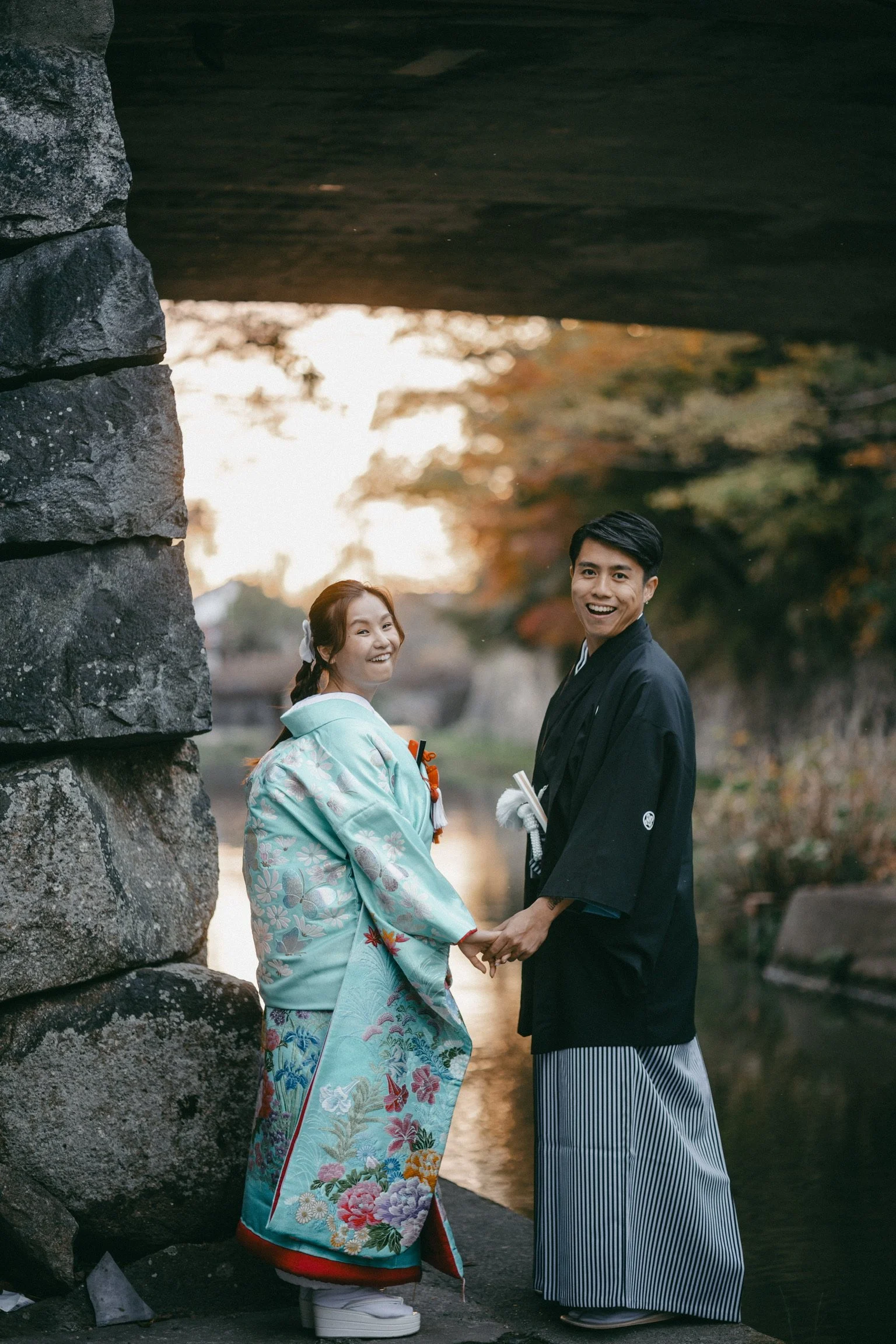 A happy couple in traditional Japanese wedding attire holding hands under a stone bridge, smiling, with a river and autumn trees in the background.