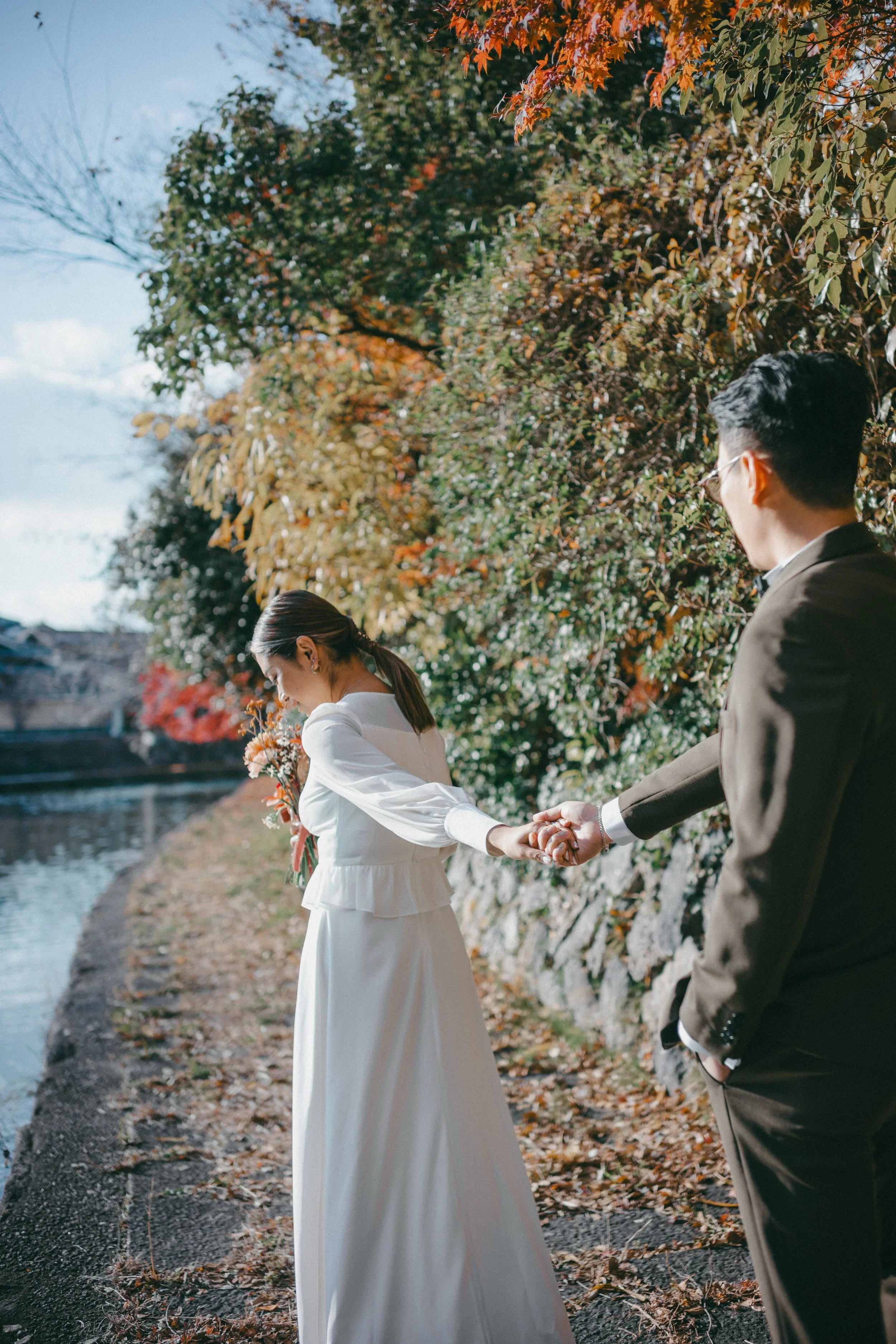 A couple holding hands outdoors near a body of water on a fall day, with autumn leaves on trees in the background.
