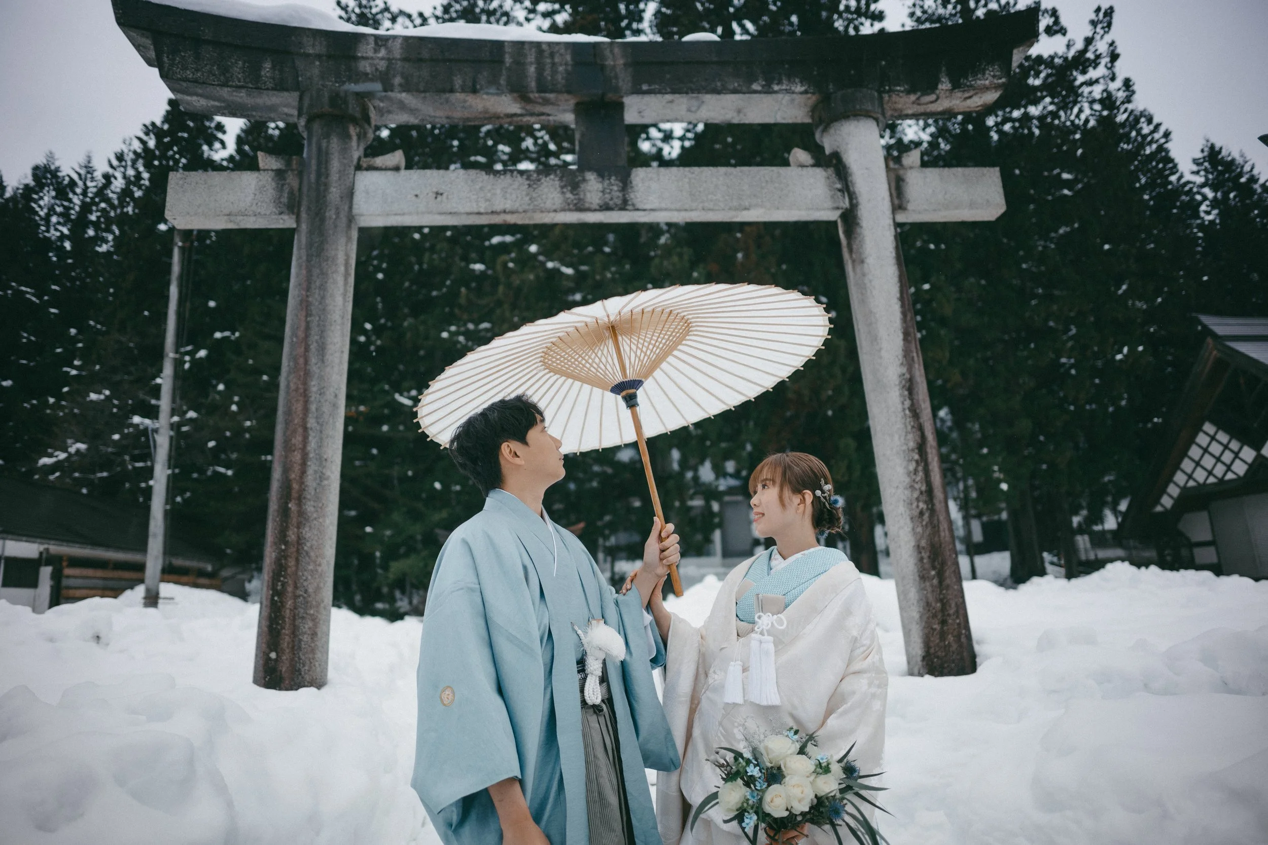 A couple dressed in traditional Japanese wedding attire stands under a large cream-colored parasol in snowy surroundings near a Shinto shrine gate. The woman holds a bouquet of white roses.
